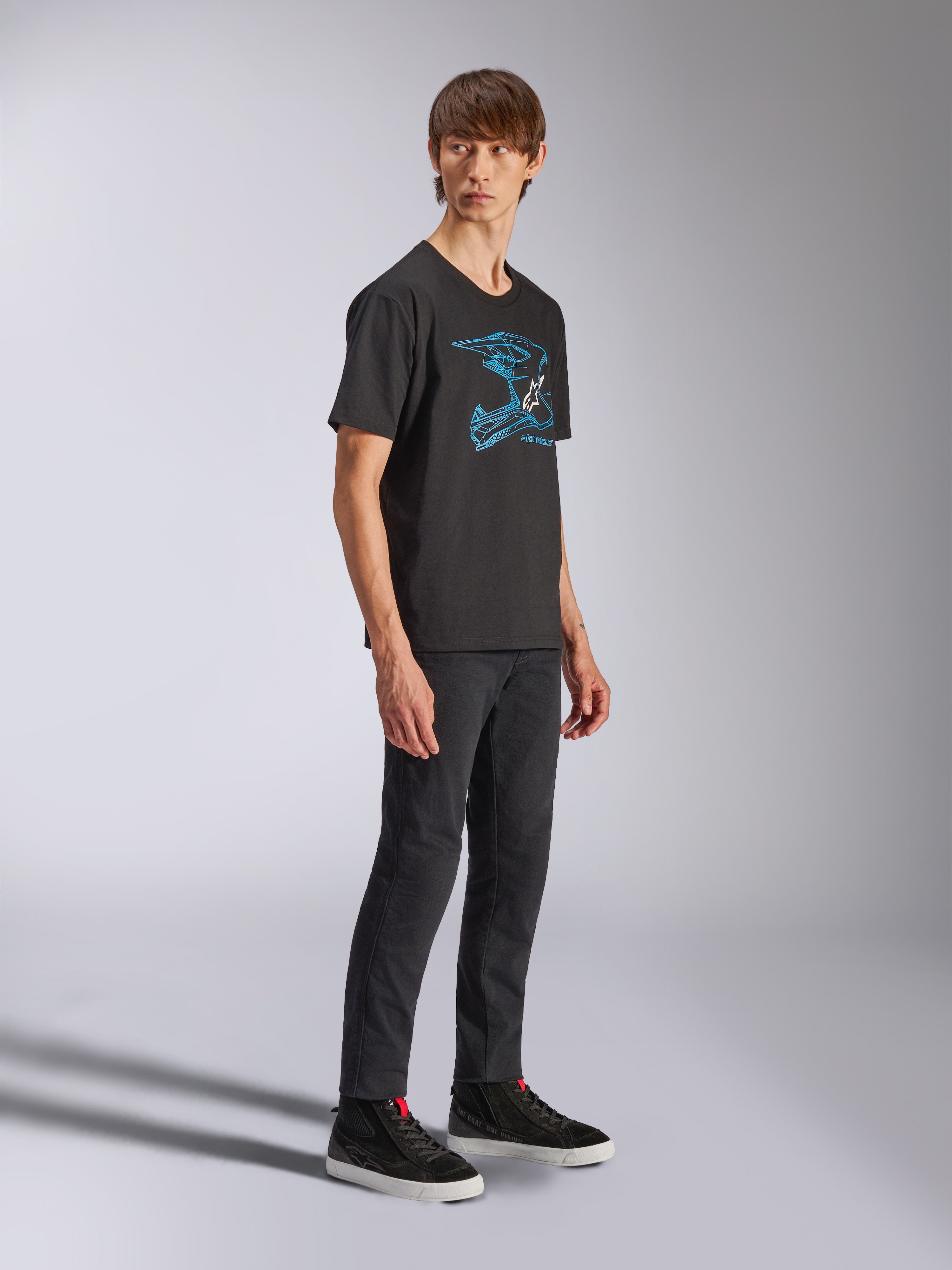 Alpinestars CSF Tee, t-shirt a maniche corte nera con grafica blu del casco MX, indossata da un modello con pantaloni neri e sneakers alte, abbigliamento casual streetwear motorsport