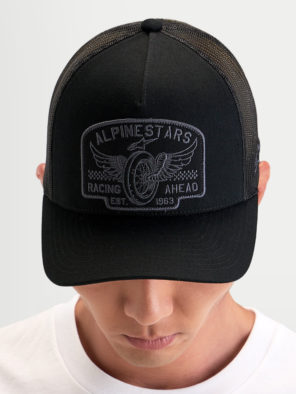 Alpinestars Cappello Heritage Patch, cappello trucker casual nero e grigio, con visiera curva, corona strutturata, retro in rete di poliestere e grande patch con logo ricamato, indossato da una persona