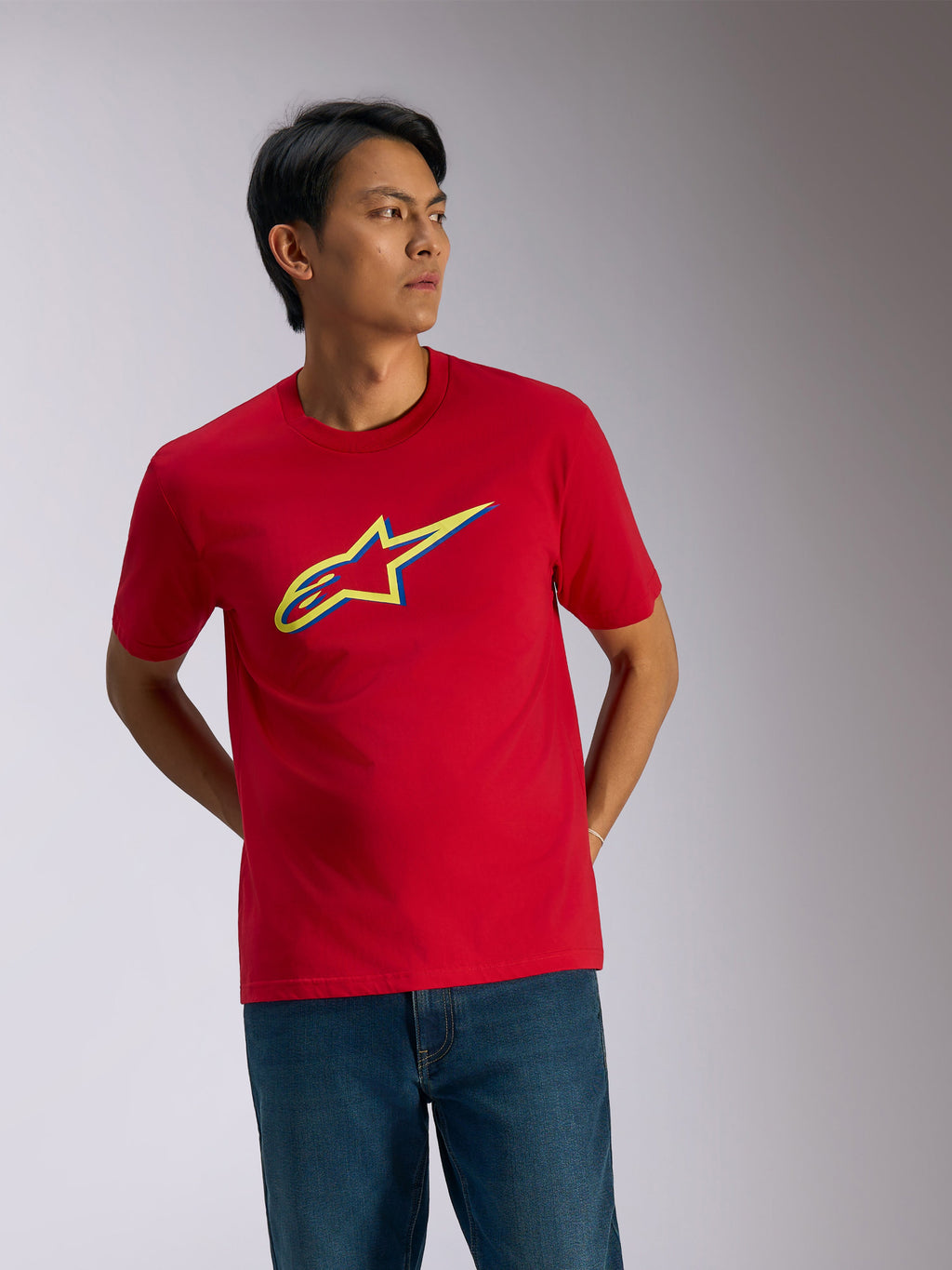 Alpinestars Ageless Shadow CSF Tee, maglietta a maniche corte, rossa, logo shadow giallo e blu sul petto, streetwear casual motorsport indossata con jeans blu