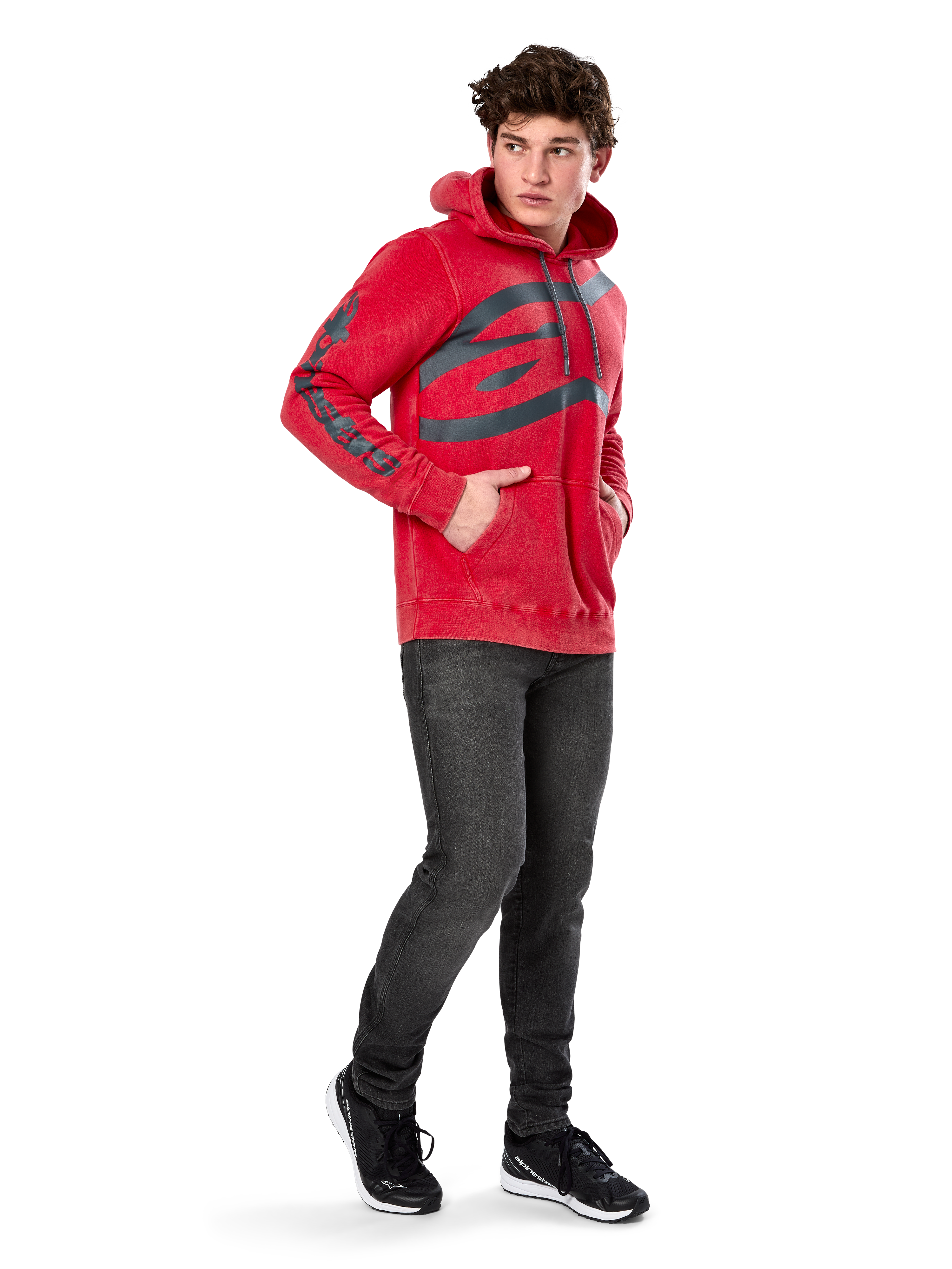 Alpinestars Unbound Hoodie, felpa in pile rossa tinta in capo, logo scuro extra large sul petto e scritta sulle maniche, tasca a marsupio, cappuccio con cordino, indossata da un modello con jeans grigio scuro e sneakers nere, streetwear casual ispirato al motorsport