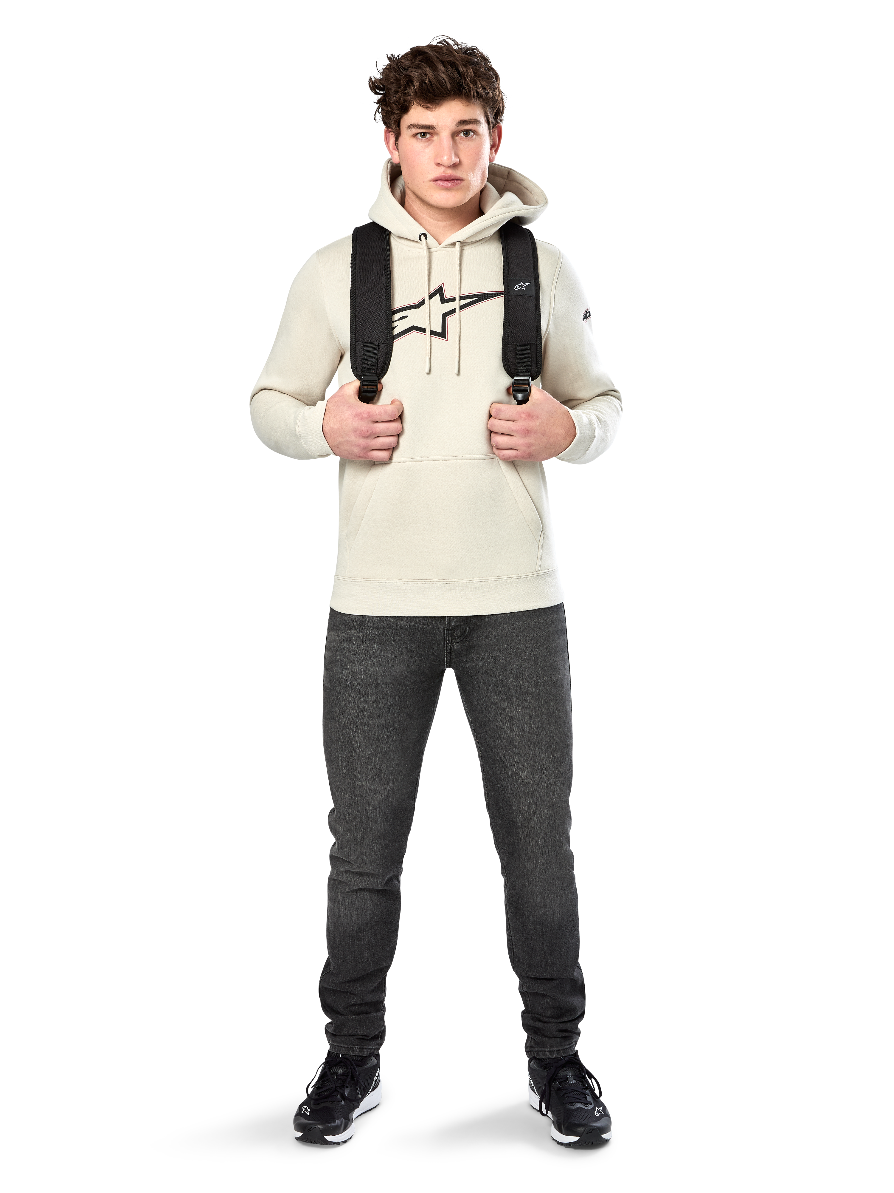 Felpa con cappuccio Alpinestars Insignia, streetwear casual, pullover in pile Off White, logo effetto fibra di carbonio con bordo rosso, cappuccio con cordino, tasca a marsupio, indossata da un modello con zaino nero e jeans grigio scuro
