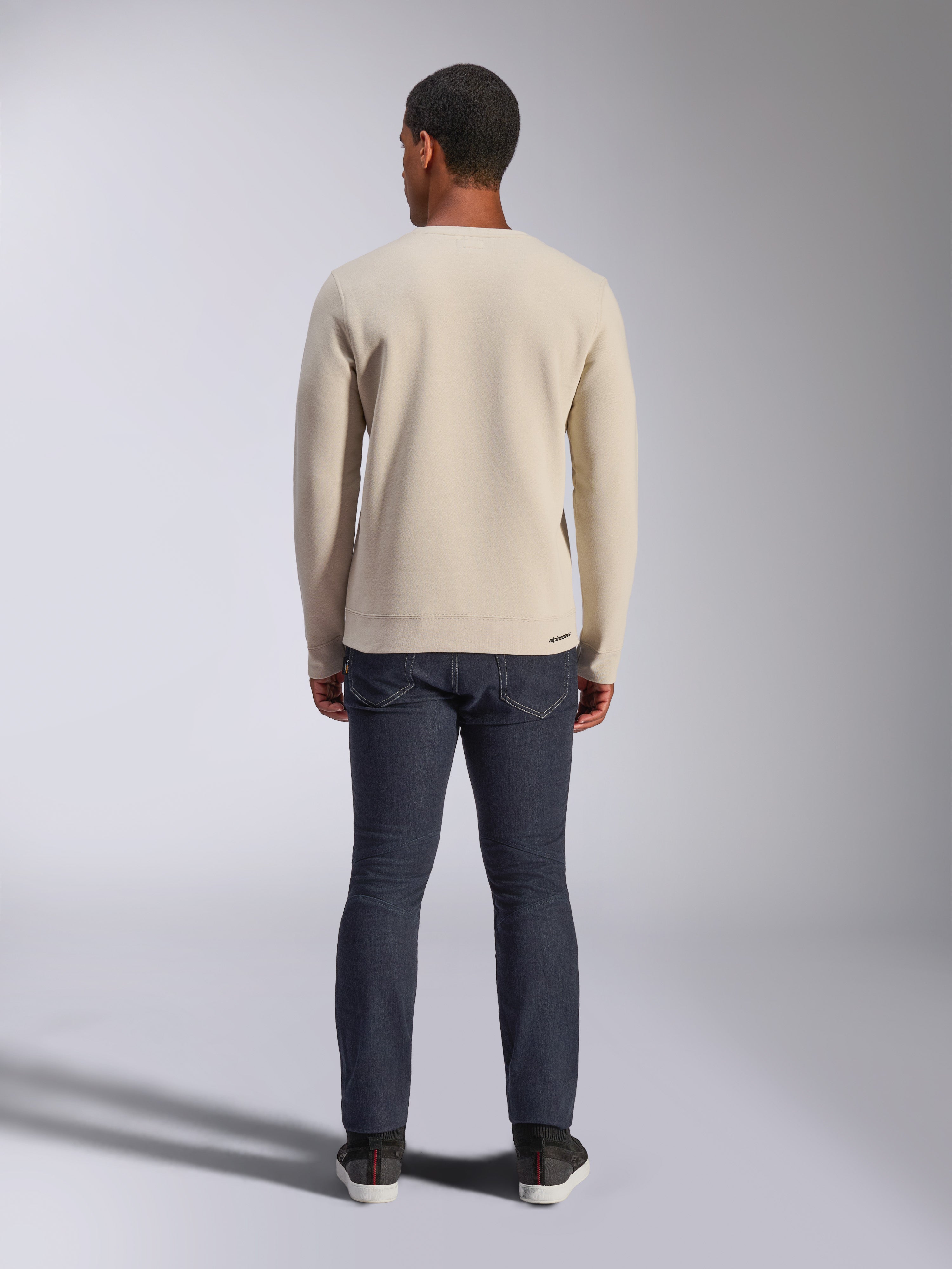 Felpa Alpinestars Positive Crew, Off White, vista posteriore di un uomo che indossa un pullover girocollo beige con bordo e polsini a coste, abbinato a jeans scuri, streetwear casual d'ispirazione motorsport.