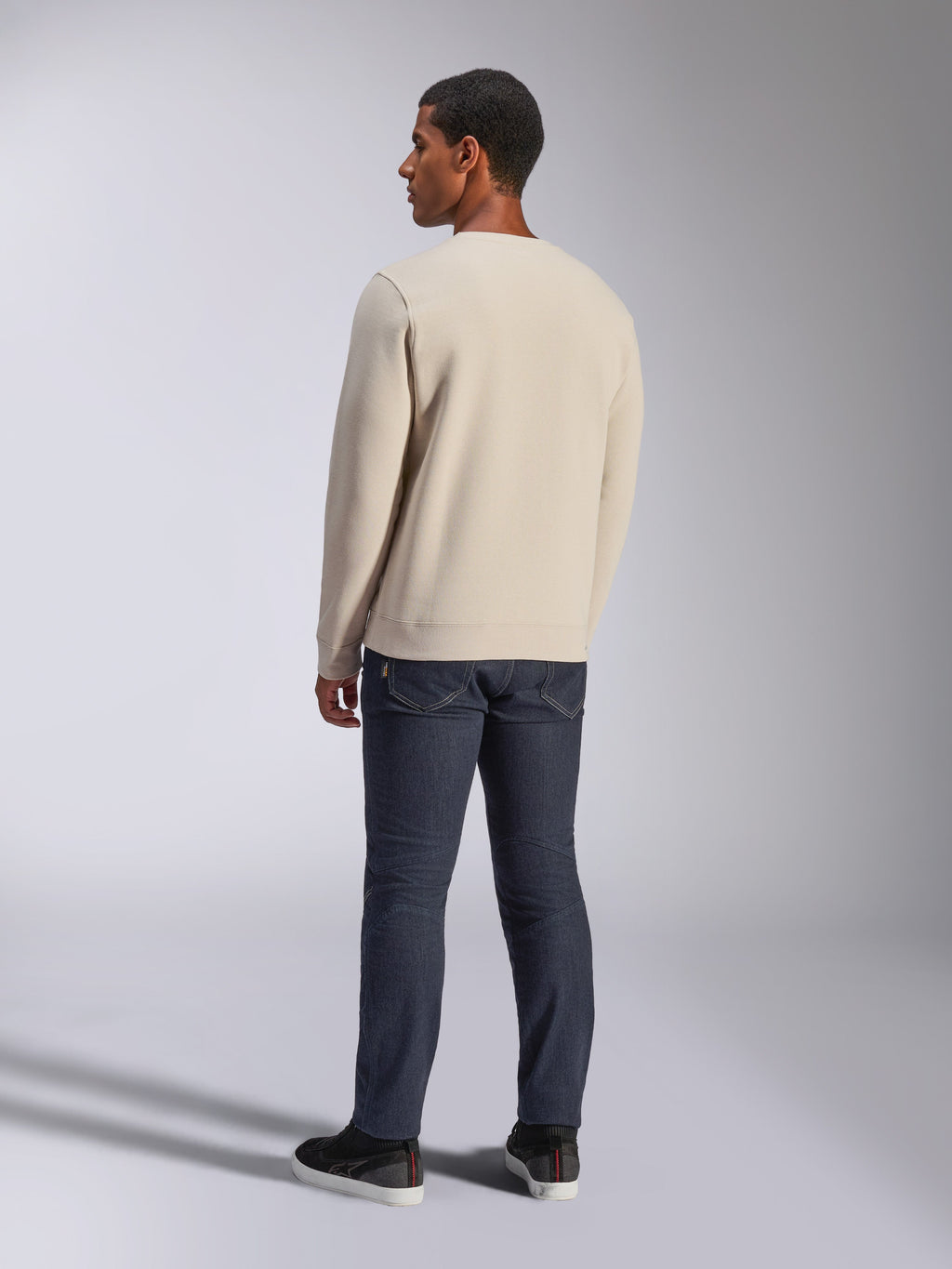 Alpinestars Positive Crew, felpa girocollo Off White, vista posteriore su modello, abbinata a jeans in denim scuro e sneakers nere, streetwear casual motorsport, con polsini e orlo a coste