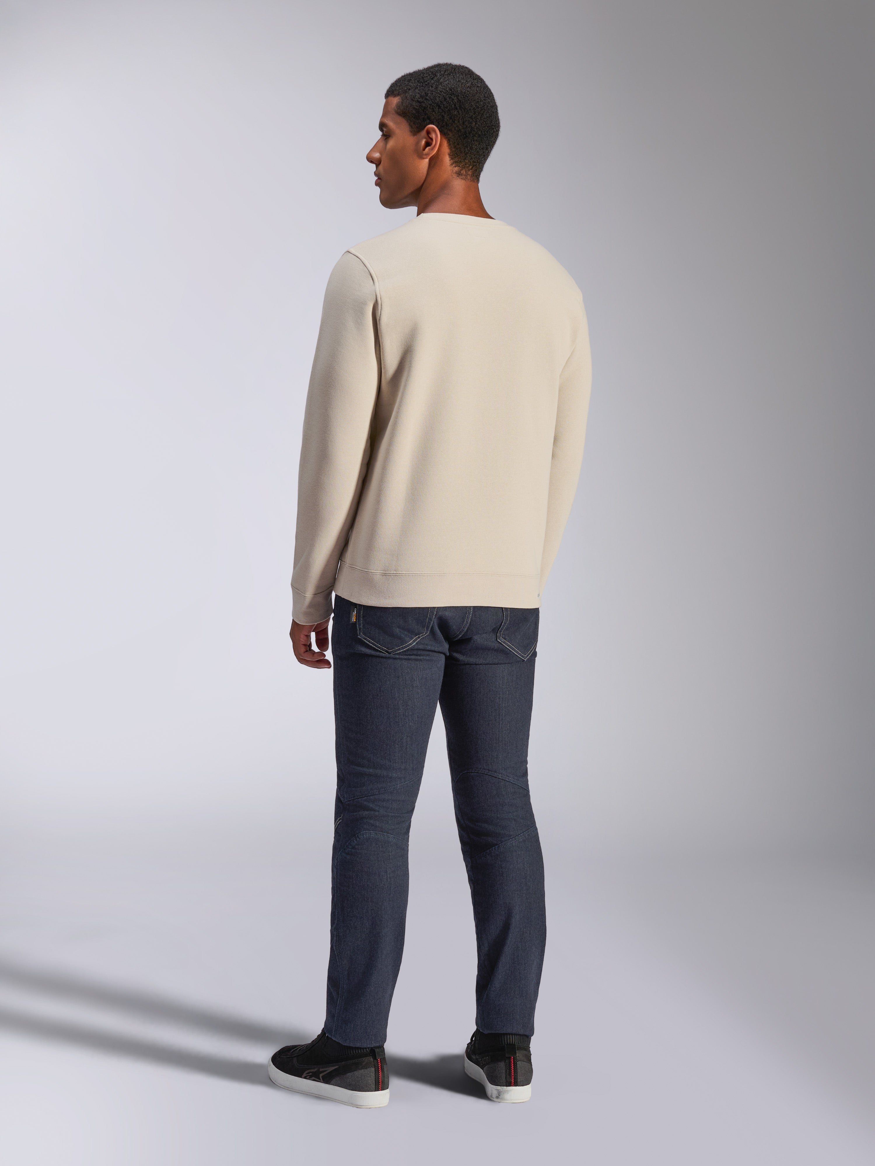 Alpinestars Positive Crew, felpa girocollo Off White, vista posteriore su modello, abbinata a jeans in denim scuro e sneakers nere, streetwear casual motorsport, con polsini e orlo a coste