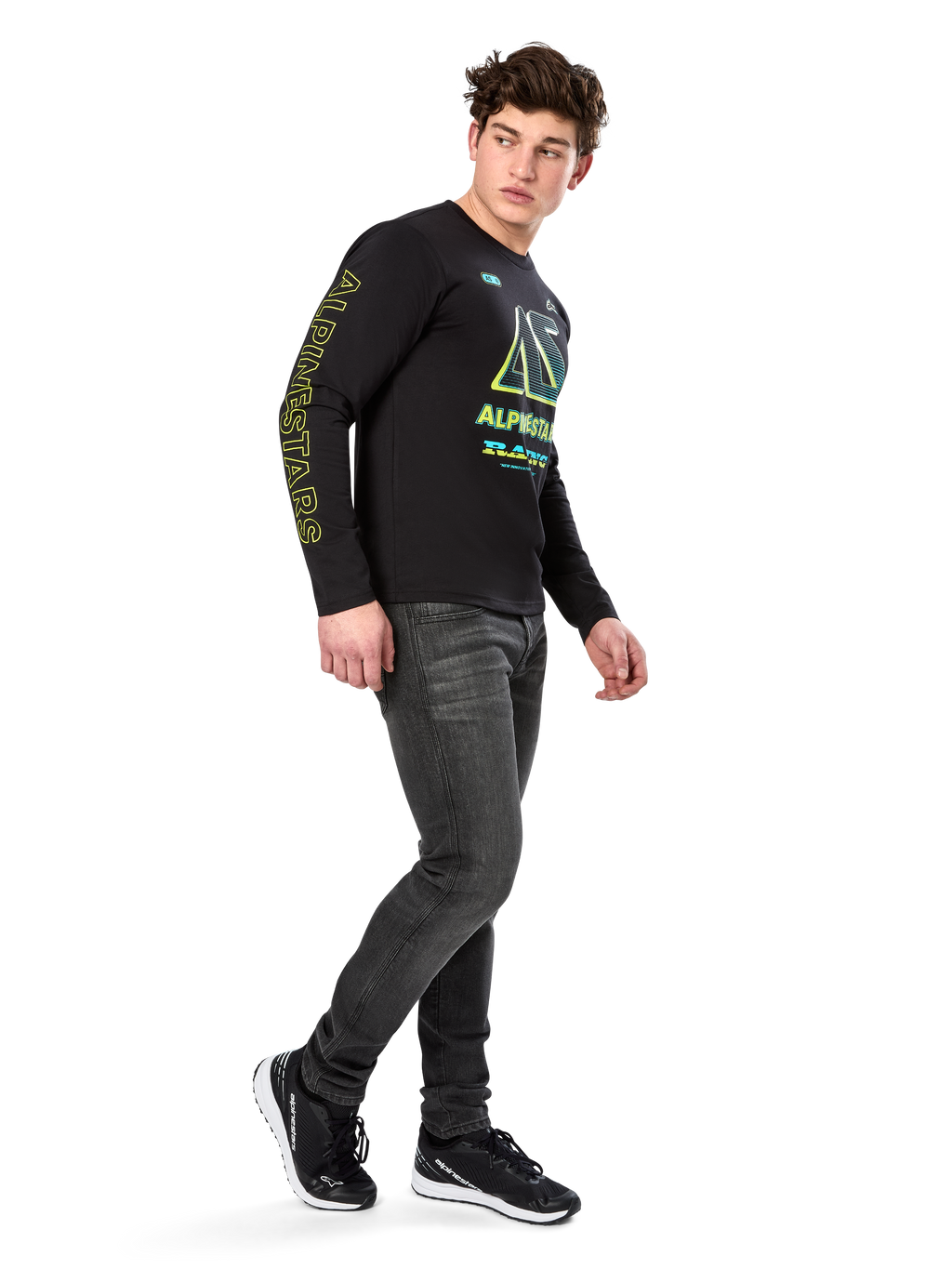 Uomo che indossa la maglietta a maniche lunghe Alpinestars Ayess CSF, nera, con scritte gialle sulle maniche e grafiche vivaci lime e ciano sul petto, abbinata a jeans grigio scuro e sneakers nere, streetwear casual ispirato al motorsport.