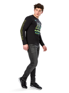Uomo che indossa la maglietta a maniche lunghe Alpinestars Ayess CSF, nera, con scritte gialle sulle maniche e grafiche vivaci lime e ciano sul petto, abbinata a jeans grigio scuro e sneakers nere, streetwear casual ispirato al motorsport.