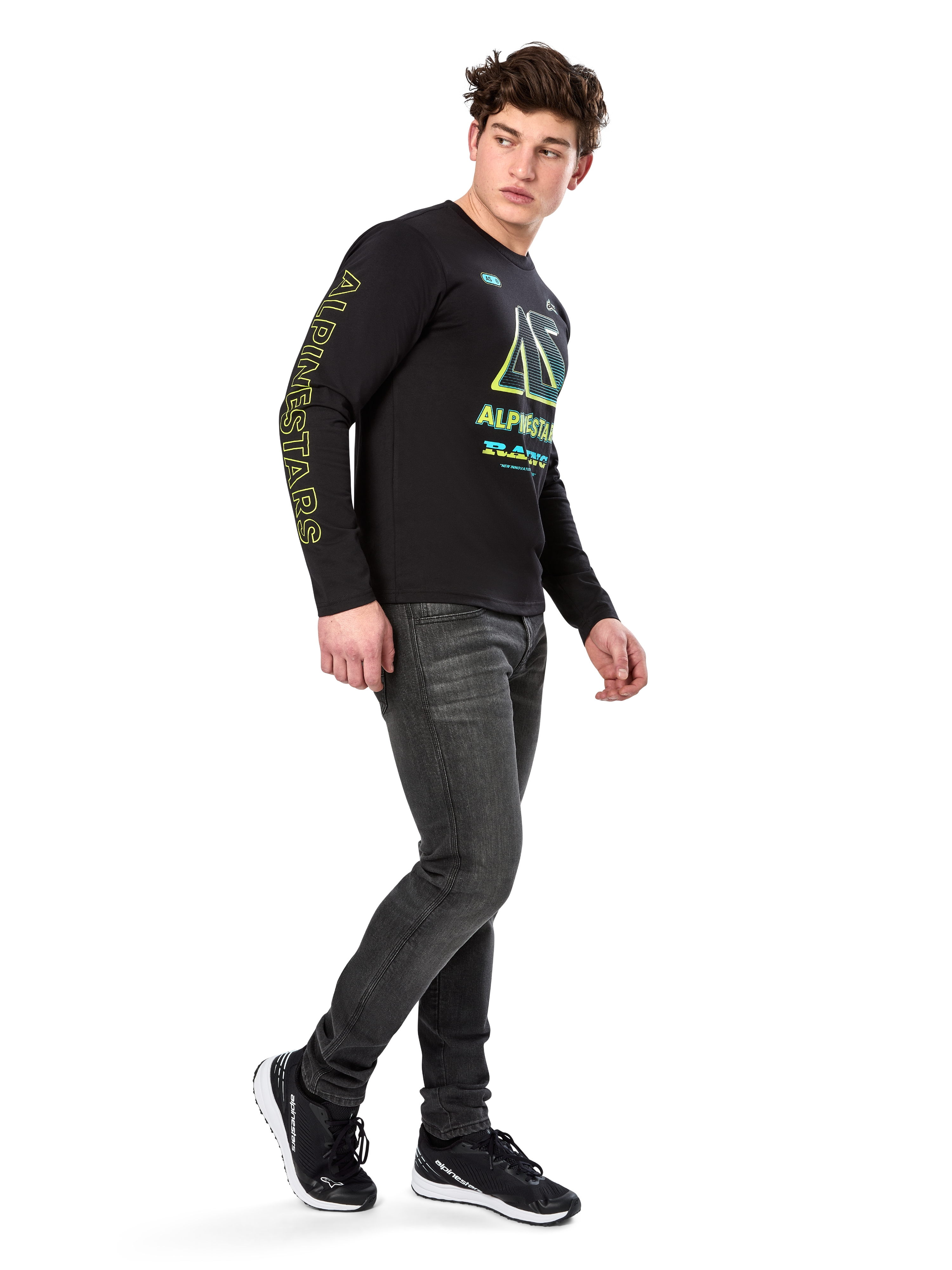 Uomo che indossa la maglietta a maniche lunghe Alpinestars Ayess CSF, nera, con scritte gialle sulle maniche e grafiche vivaci lime e ciano sul petto, abbinata a jeans grigio scuro e sneakers nere, streetwear casual ispirato al motorsport.