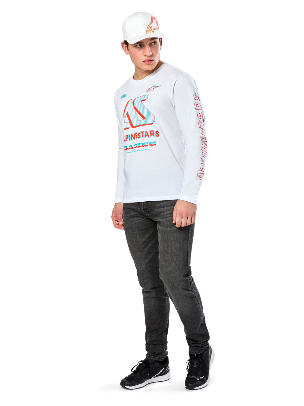 T-shirt Alpinestars Ayess CSF, maglietta a maniche lunghe, bianca, con grandi grafiche racing CSF blu e rosse sul petto e marchio Alpinestars rosso sulla manica, indossata con jeans scuri e un cappellino Alpinestars bianco, streetwear casual motorsport