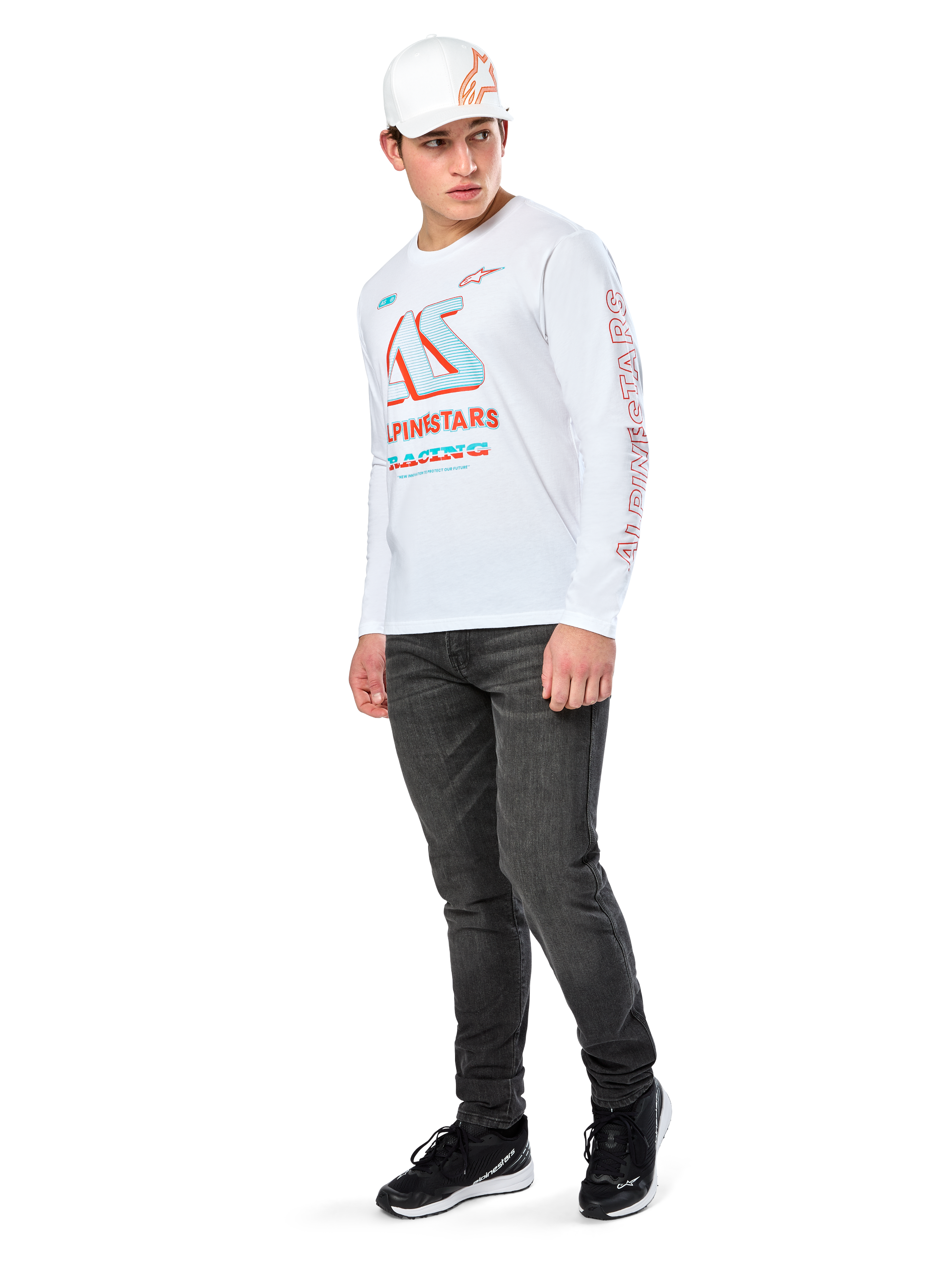 T-shirt Alpinestars Ayess CSF, maglietta a maniche lunghe, bianca, con grandi grafiche racing CSF blu e rosse sul petto e marchio Alpinestars rosso sulla manica, indossata con jeans scuri e un cappellino Alpinestars bianco, streetwear casual motorsport