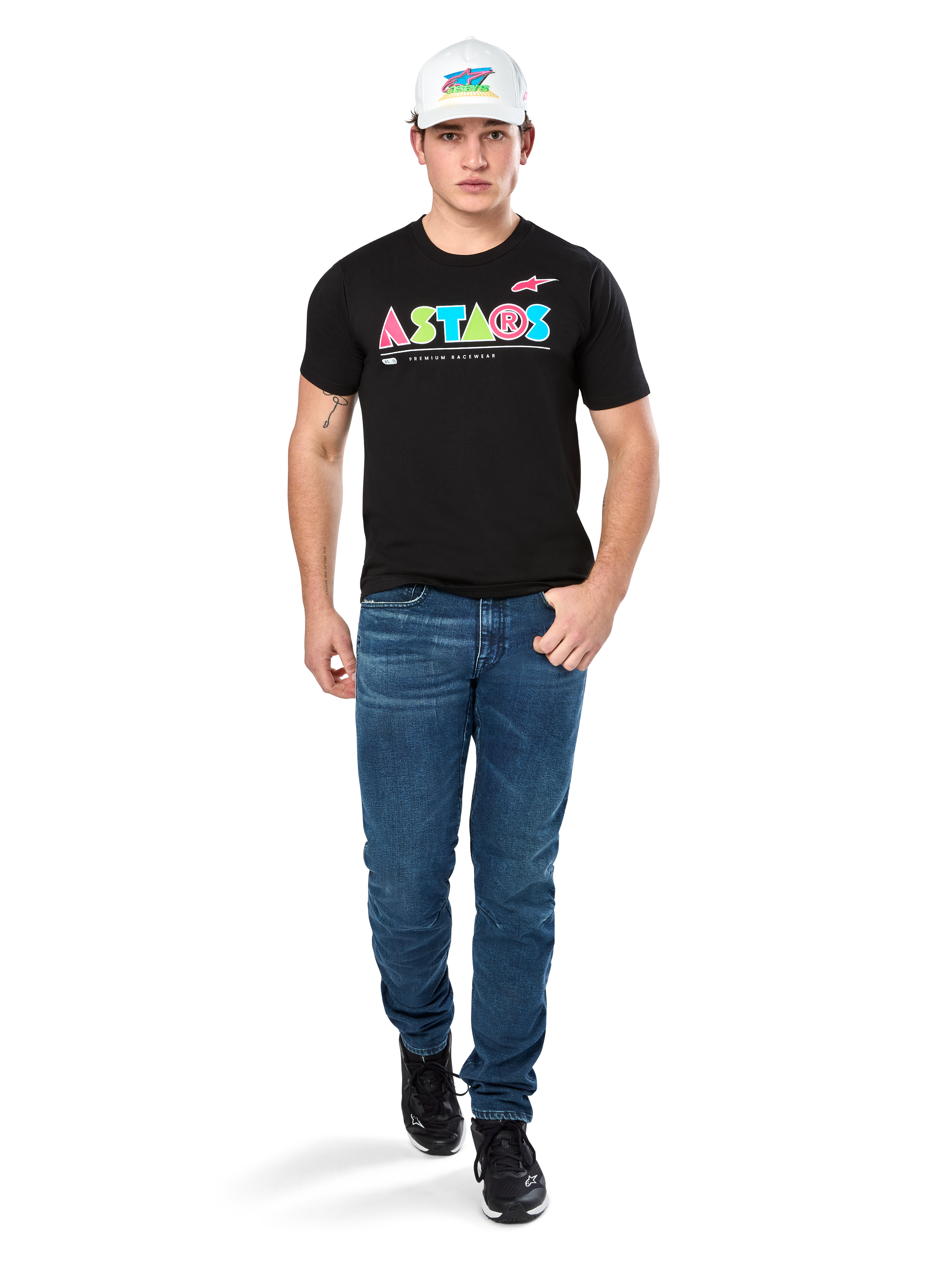 Modello maschile che indossa la maglietta Alpinestars Cereal CSF, T-shirt nera a maniche corte con logo ASTARS geometrico colorato, abbinata a jeans blu e cappellino bianco, streetwear casual ispirato al motorsport