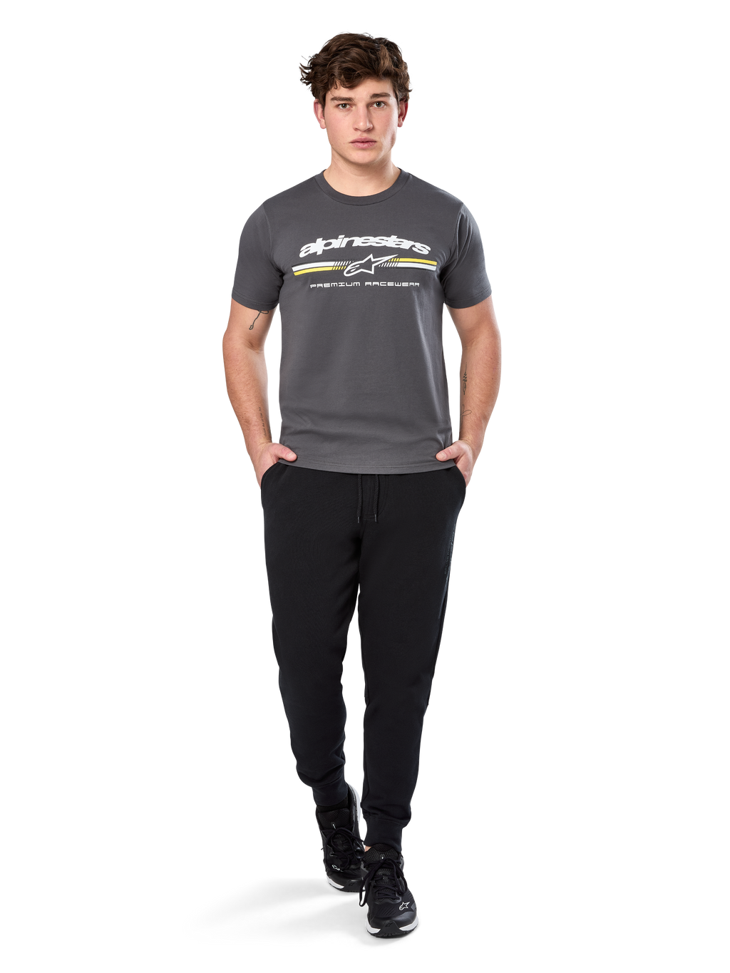 Uomo che indossa la Alpinestars Prevail Csf Tee, t-shirt a maniche corte, grigio antracite, con logo Alpinestars bianco e striscia tricolore con accenti gialli, abbinata a pantaloni sportivi neri e sneakers, abbigliamento casual motorsport streetwear