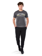 Uomo che indossa la Alpinestars Prevail Csf Tee, t-shirt a maniche corte, grigio antracite, con logo Alpinestars bianco e striscia tricolore con accenti gialli, abbinata a pantaloni sportivi neri e sneakers, abbigliamento casual motorsport streetwear