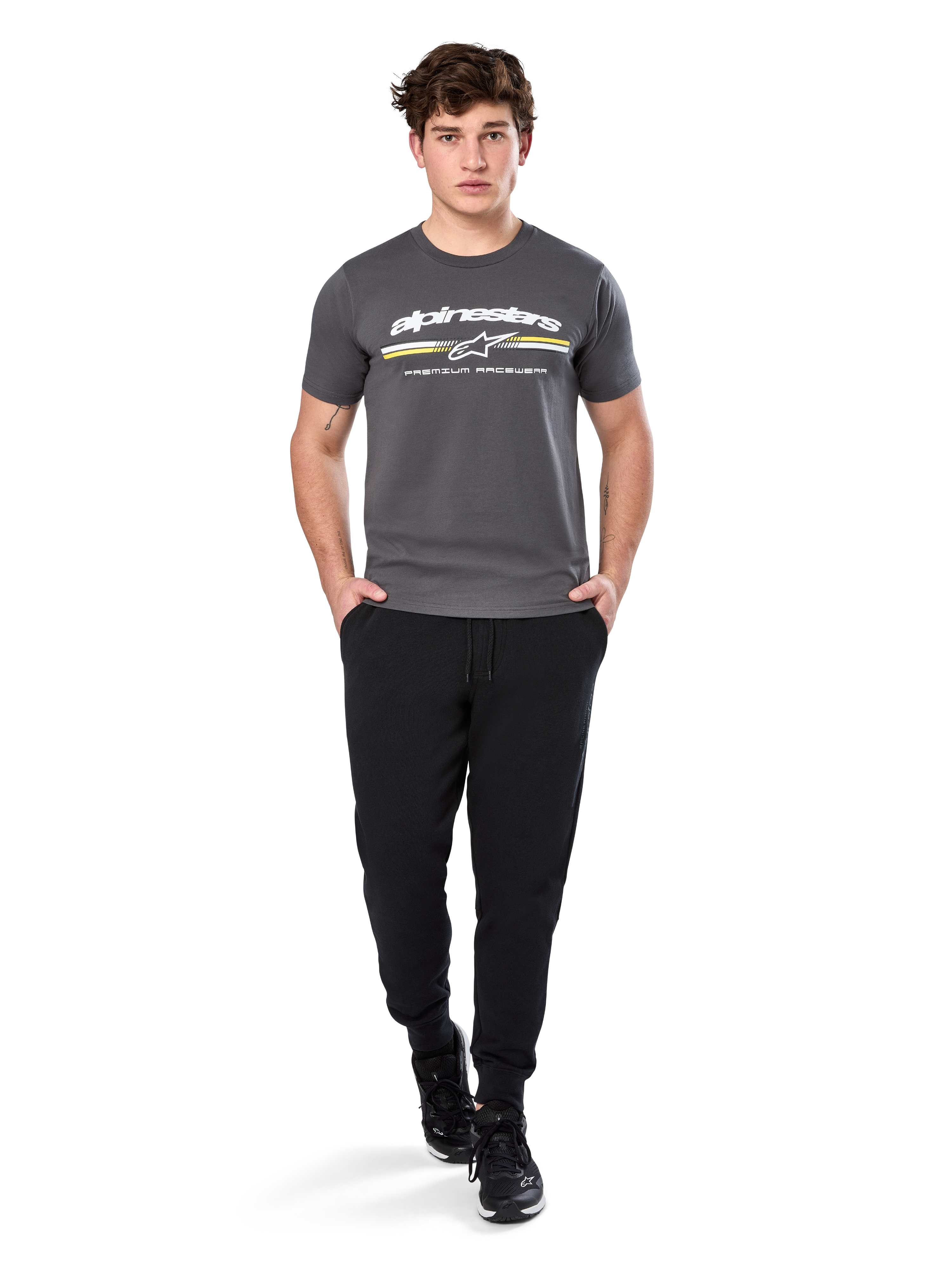Uomo che indossa la Alpinestars Prevail Csf Tee, t-shirt a maniche corte, grigio antracite, con logo Alpinestars bianco e striscia tricolore con accenti gialli, abbinata a pantaloni sportivi neri e sneakers, abbigliamento casual motorsport streetwear