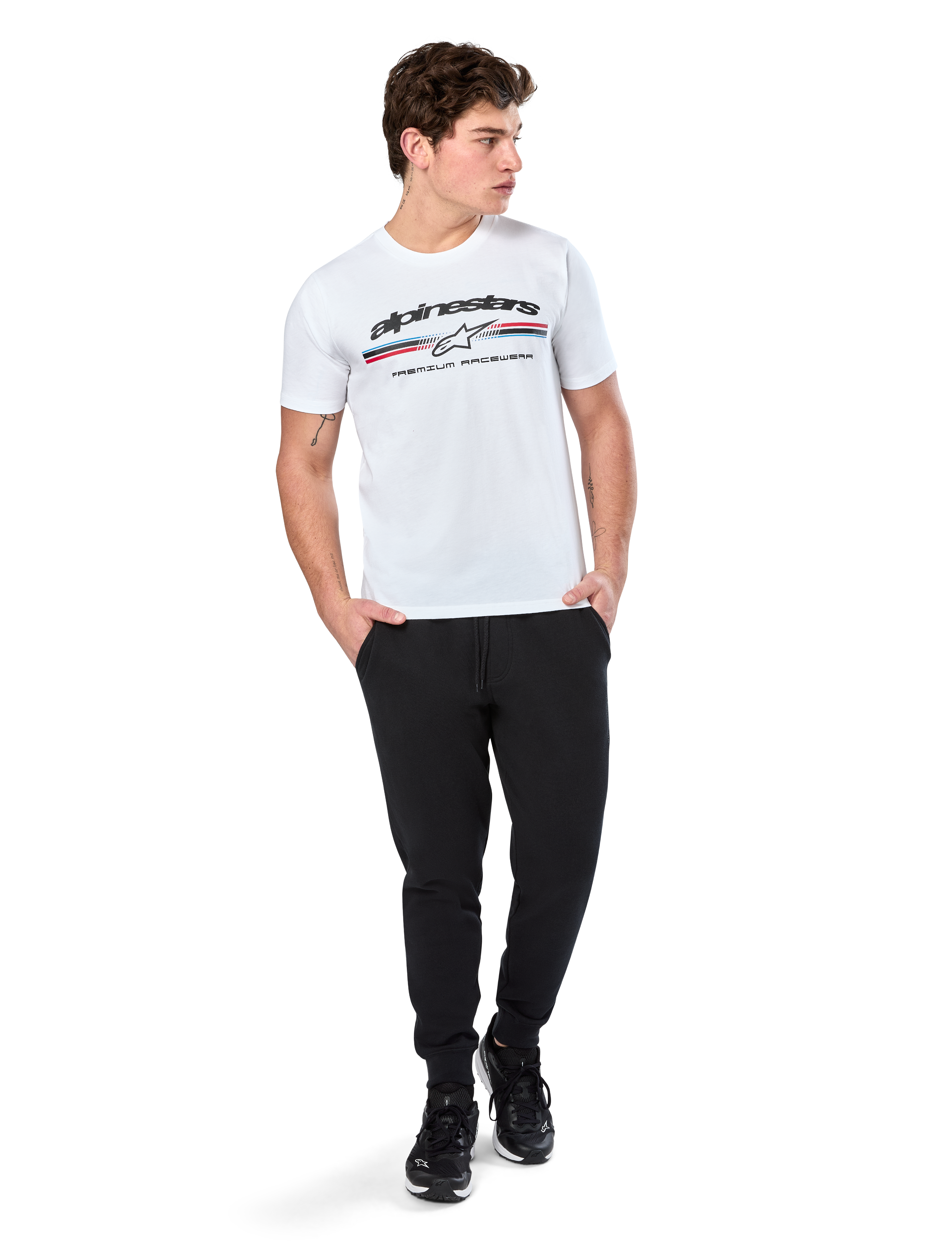 Alpinestars T-shirt Prevail CSF, t-shirt a maniche corte, bianca, con logo a strisce tricolore stile paddock anteriore e marchio Alpinestars, indossata da un modello con pantaloni sportivi neri e sneaker nere, streetwear casual da motorsport
