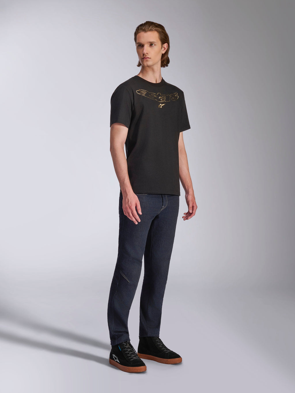 T-shirt Alpinestars Luxo CSF, maglietta a maniche corte, nera, logo Astars dorato profilato sul petto, indossata da un modello con jeans denim scuro e sneakers nere, streetwear casual motorsport