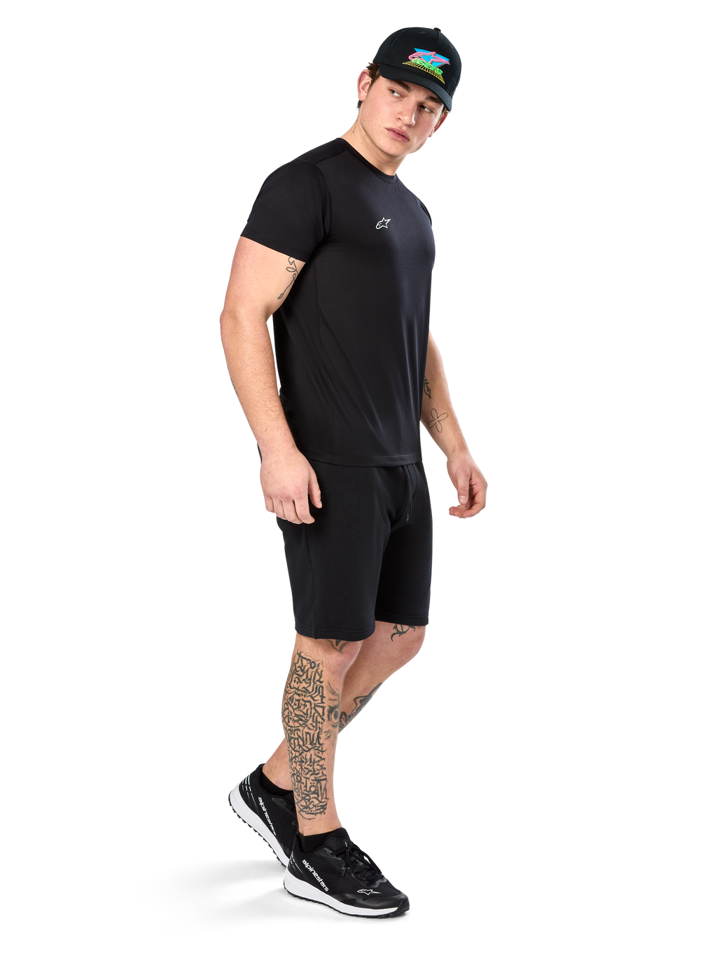 Uomo con tatuaggi che indossa Alpinestars Point Performance Tee, t-shirt tecnica nera a maniche corte, piccolo logo bianco, abbinata a pantaloncini neri, cappellino nero con grafica colorata e sneakers bianche e nere, stile casual sportivo