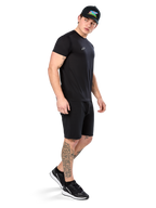 Uomo con tatuaggi che indossa Alpinestars Point Performance Tee, t-shirt tecnica nera a maniche corte, piccolo logo bianco, abbinata a pantaloncini neri, cappellino nero con grafica colorata e sneakers bianche e nere, stile casual sportivo
