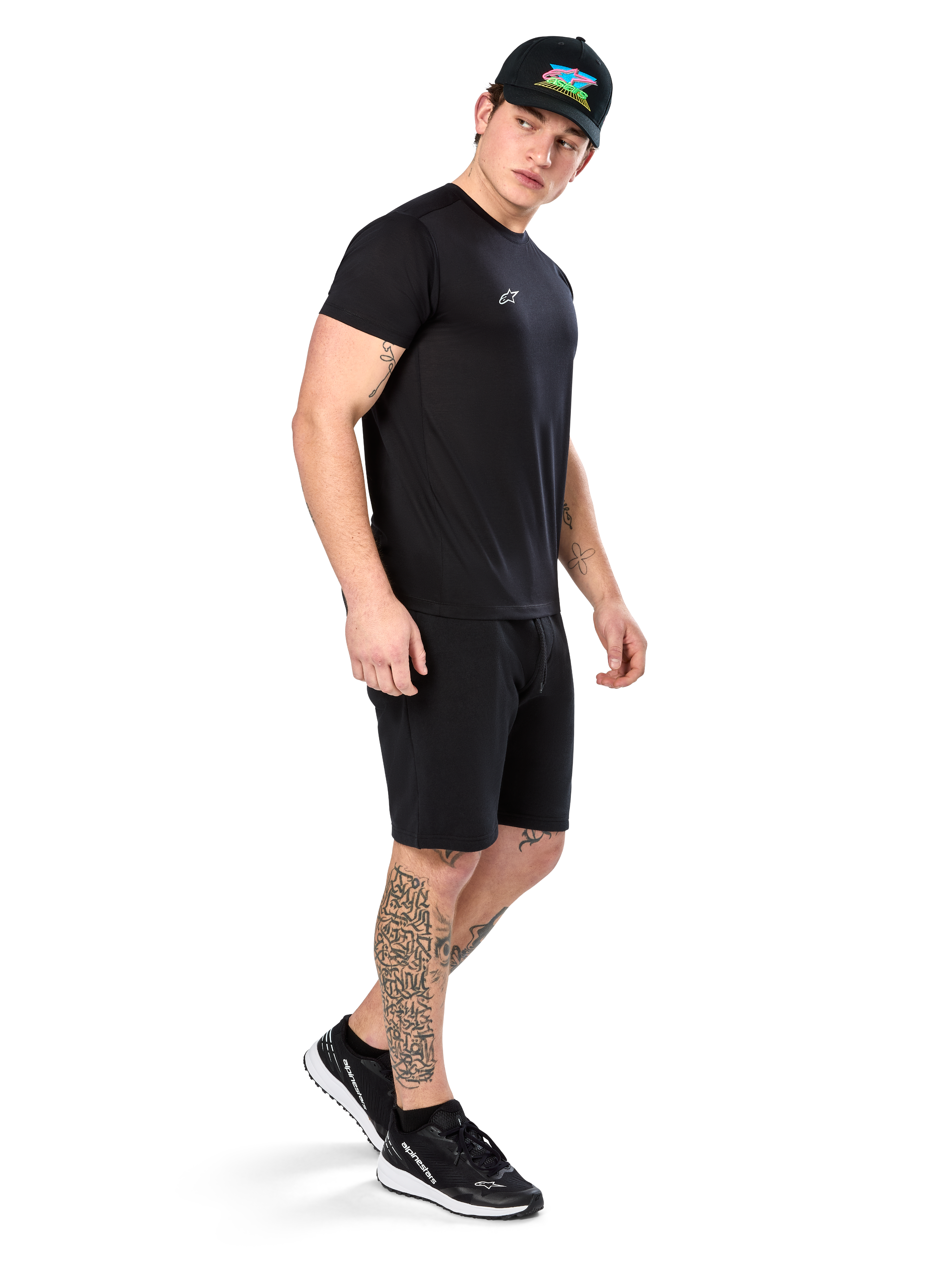 Uomo con tatuaggi che indossa Alpinestars Point Performance Tee, t-shirt tecnica nera a maniche corte, piccolo logo bianco, abbinata a pantaloncini neri, cappellino nero con grafica colorata e sneakers bianche e nere, stile casual sportivo