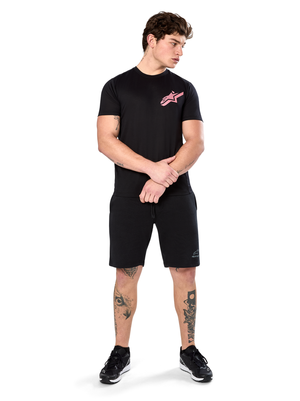 Modello che indossa Alpinestars Attrition Performance Tee, t-shirt performance a maniche corte, nera con logo Astars rosa sul petto, abbinata a pantaloncini e sneakers neri, abbigliamento sportivo casual ispirato al motorsport