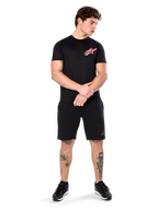 Modello che indossa Alpinestars Attrition Performance Tee, t-shirt performance a maniche corte, nera con logo Astars rosa sul petto, abbinata a pantaloncini e sneakers neri, abbigliamento sportivo casual ispirato al motorsport