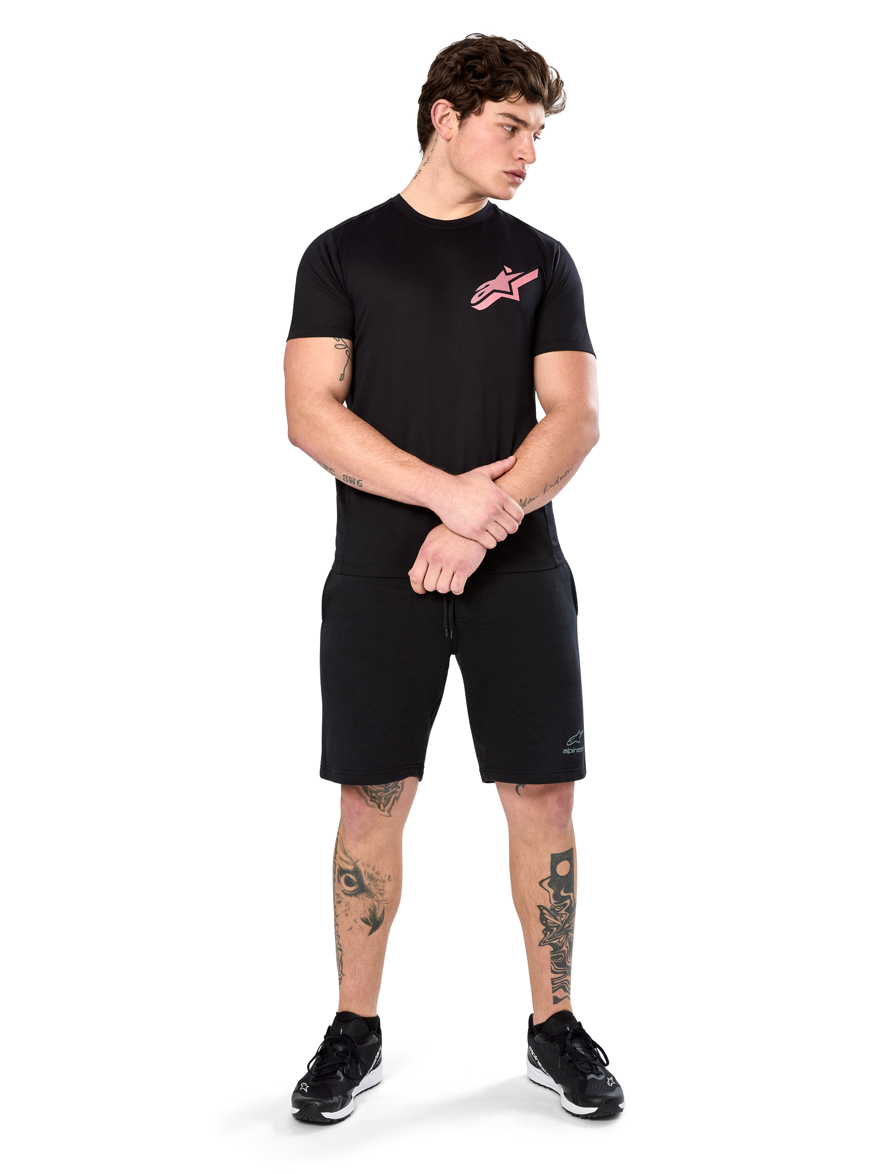 Modello che indossa Alpinestars Attrition Performance Tee, t-shirt performance a maniche corte, nera con logo Astars rosa sul petto, abbinata a pantaloncini e sneakers neri, abbigliamento sportivo casual ispirato al motorsport