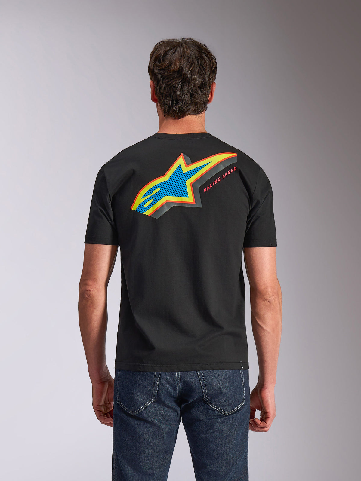Alpinestars Angler CSF Tee, t-shirt a maniche corte, nero, vista posteriore con vivace logo Astars multicolore con riempimento traforato e scritta 'RACING AHEAD', indossata con jeans blu, abbigliamento casual ispirato al motorsport