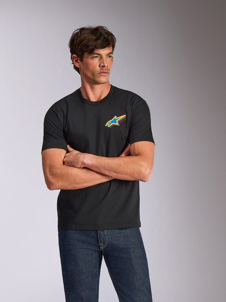 Alpinestars Angler CSF Tee, maglietta a maniche corte, nero, con un vivace logo Astars multicolore sul petto, indossata da un modello con le braccia incrociate e jeans blu, streetwear casual ispirato al motorsport