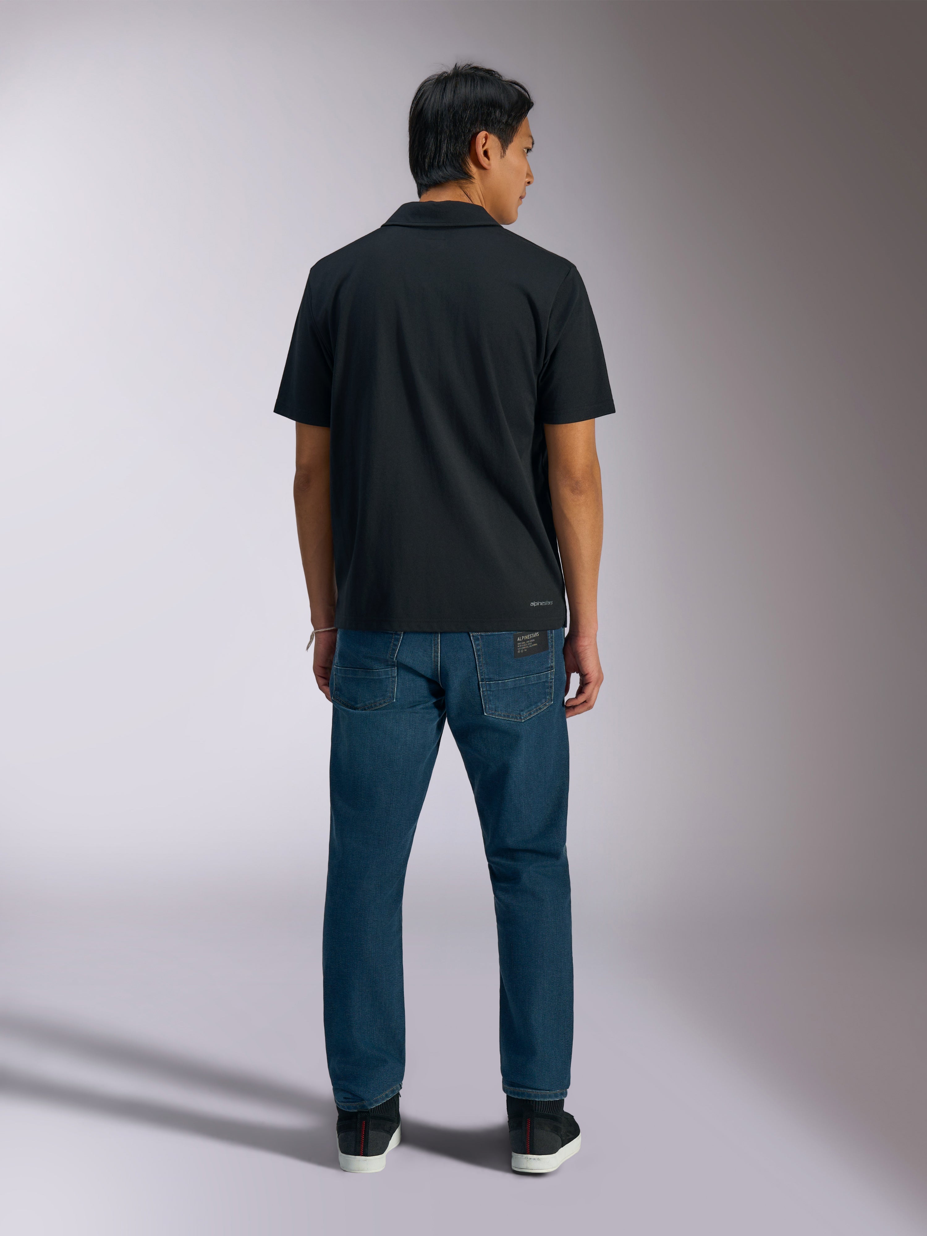 Alpinestars Impetus Sport Polo, nero, vista posteriore della polo a maniche corte con logo Alpinestars sulla parte inferiore della schiena, indossata con jeans blu, abbigliamento sportivo casual ispirato al motorsport