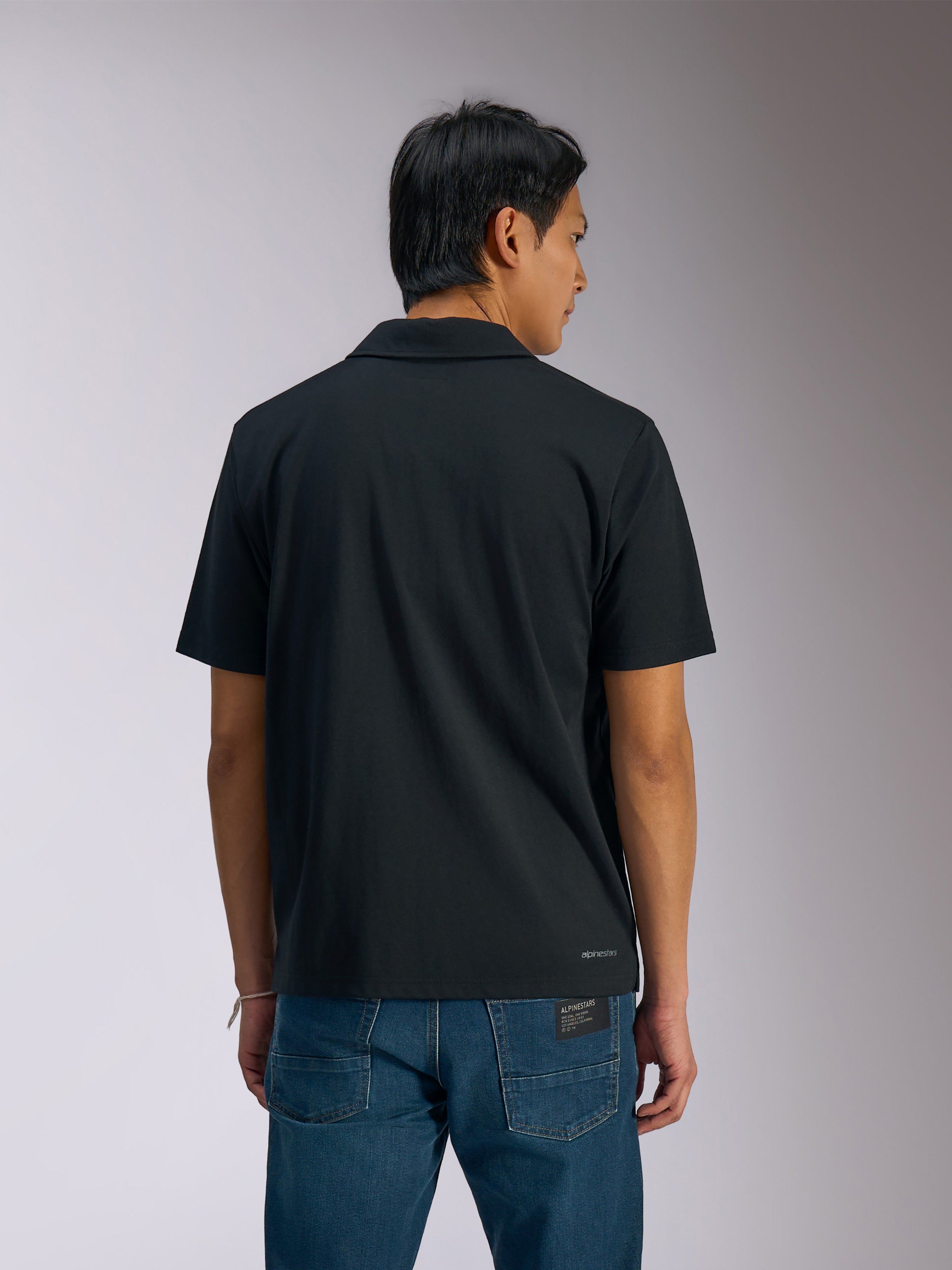 Alpinestars Impetus Sport Polo, polo a maniche corte nera, vista posteriore di un uomo che indossa la polo con jeans in denim blu, con piccola scritta Alpinestars sulla parte inferiore della schiena, abbigliamento sportivo casual ispirato al motorsport