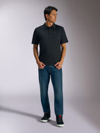 Alpinestars Impetus Sport Polo, polo nera a maniche corte, con chiusura a tre bottoni e logo Astars grigio sul petto, indossata da un uomo con jeans blu e sneakers alte nere, abbigliamento sportivo casual ispirato al motorsport