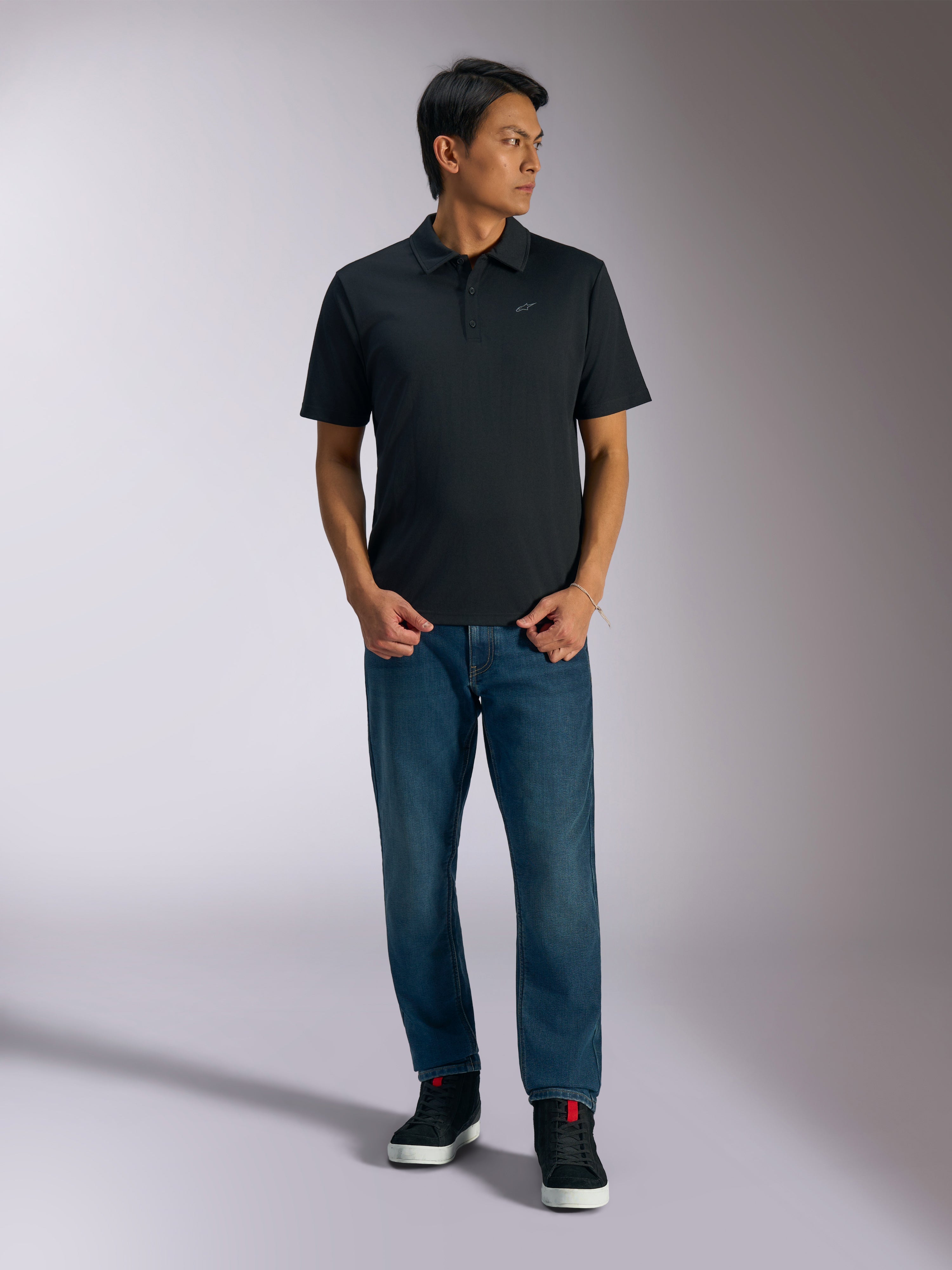 Alpinestars Impetus Sport Polo, polo nera a maniche corte, con chiusura a tre bottoni e logo Astars grigio sul petto, indossata da un uomo con jeans blu e sneakers alte nere, abbigliamento sportivo casual ispirato al motorsport