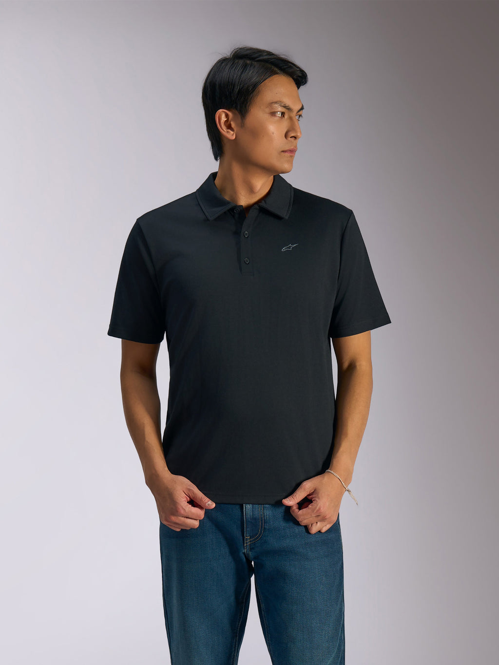 Polo Alpinestars Impetus Sport, polo a maniche corte nera, chiusura a tre bottoni e colletto dello stesso tessuto, piccolo logo Astars grigio sul petto a sinistra, indossata con jeans blu, abbigliamento sportivo casual ispirato al motorsport