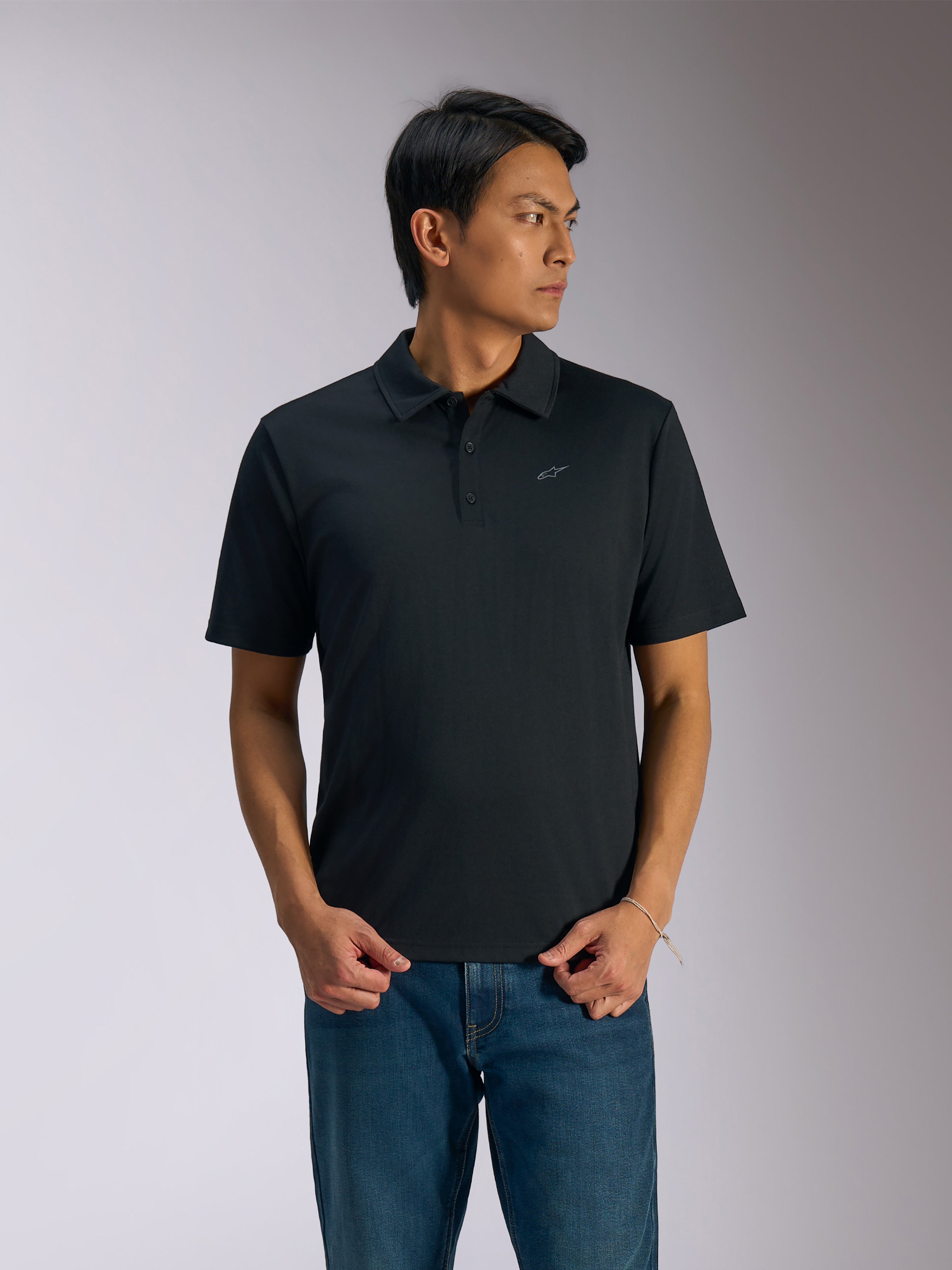 Polo Alpinestars Impetus Sport, polo a maniche corte nera, chiusura a tre bottoni e colletto dello stesso tessuto, piccolo logo Astars grigio sul petto a sinistra, indossata con jeans blu, abbigliamento sportivo casual ispirato al motorsport
