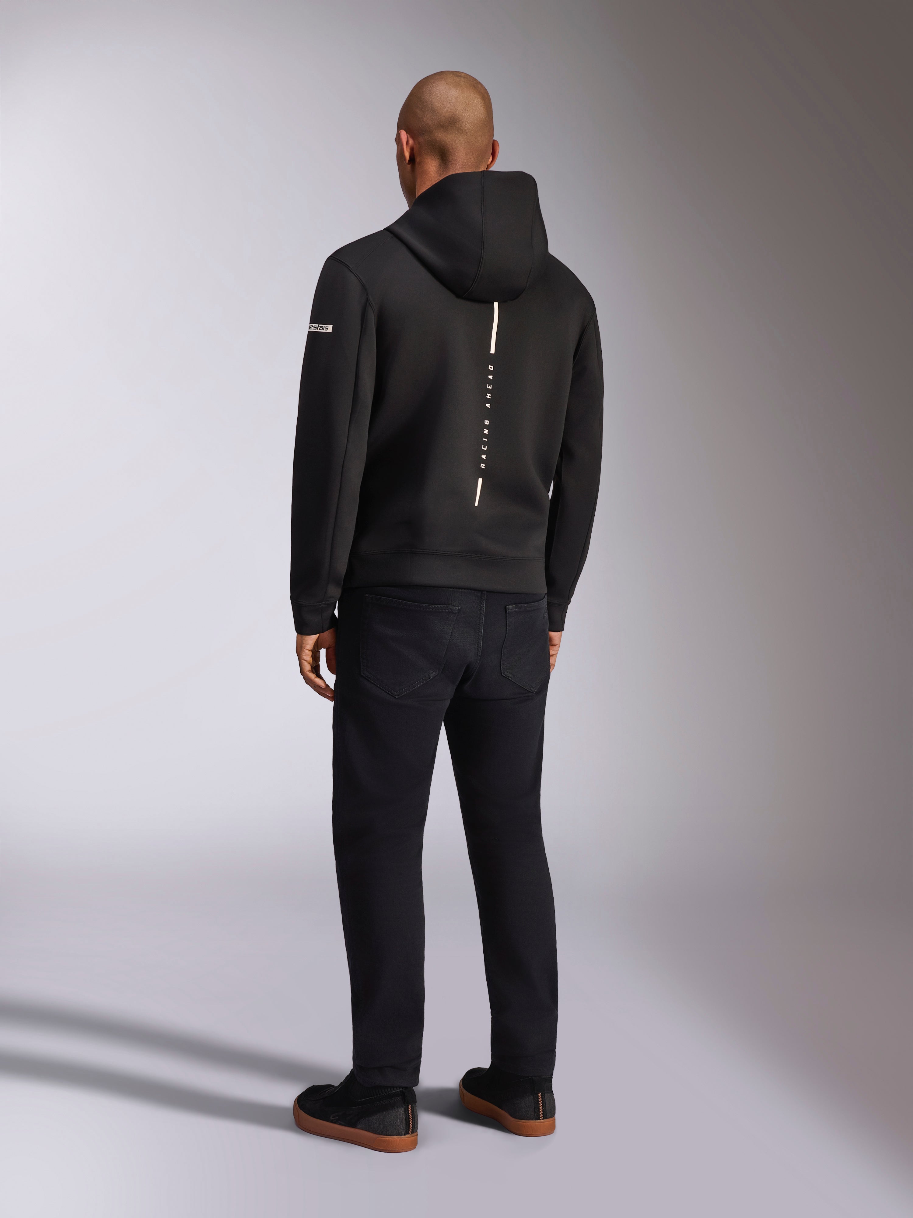 Alpinestars Insight Hoodie, felpa sportiva nera, vista posteriore con striscia bianca verticale e branding 'RACING AHEAD', logo sulla manica, abbinata a pantaloni neri e sneakers, design casual ispirato al motorsport