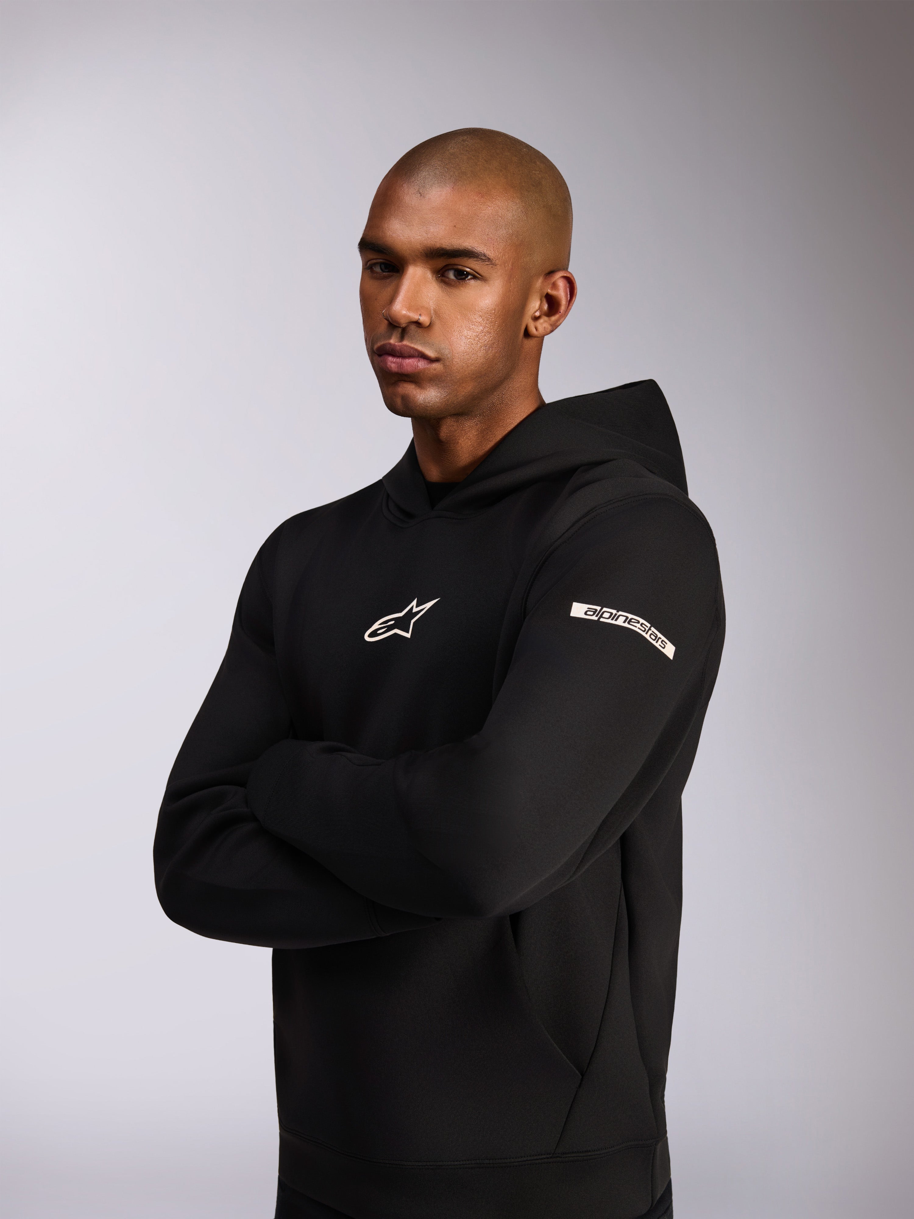 Alpinestars Insight Hoodie, felpa sportiva casual, nera, logo Astars bianco sul petto e logo Alpinestars Word sulla manica, modello che indossa la felpa con le braccia incrociate, pregiata costruzione in pile double face
