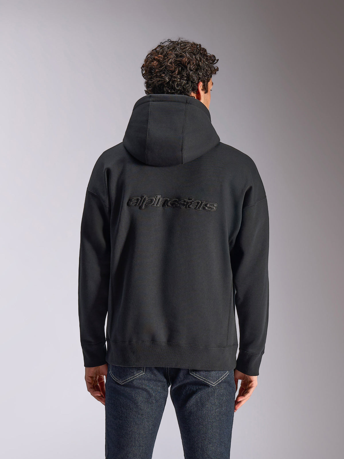 Alpinestars Emergent Oversized Hoodie, vista posteriore di una felpa nera con cappuccio indossata da un modello, con logo Alpinestars ricamato sul retro, vestibilità oversize, streetwear casual abbinato a jeans scuri