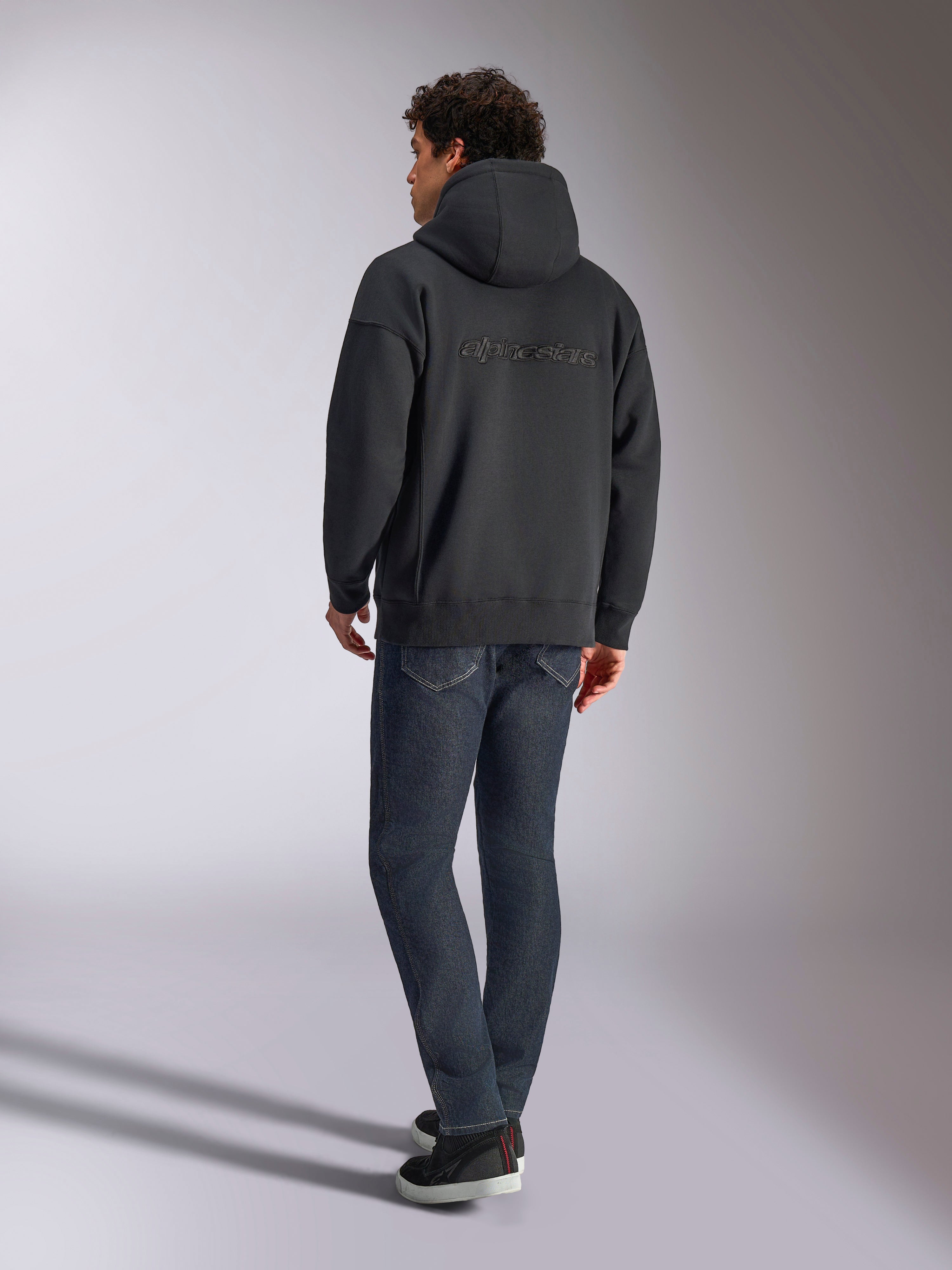 Felpa con cappuccio Alpinestars Emergent Oversized, nero, vista posteriore con logo Alpinestars in ricamo 3D sulle spalle, vestibilità oversize, abbinata a jeans in denim blu scuro e sneaker nere, streetwear casual ispirato al motorsport