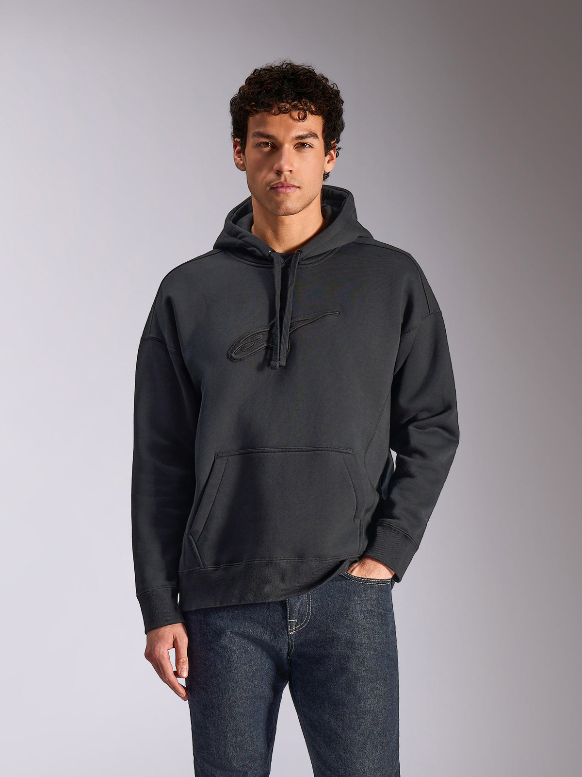 Alpinestars Emergent Oversized Hoodie, nero, modello che indossa una felpa con cappuccio oversize in pile, logo Astars ricamato in 3D tono su tono, tasca a marsupio, cappuccio con cordino, streetwear casual ispirato al motorsport