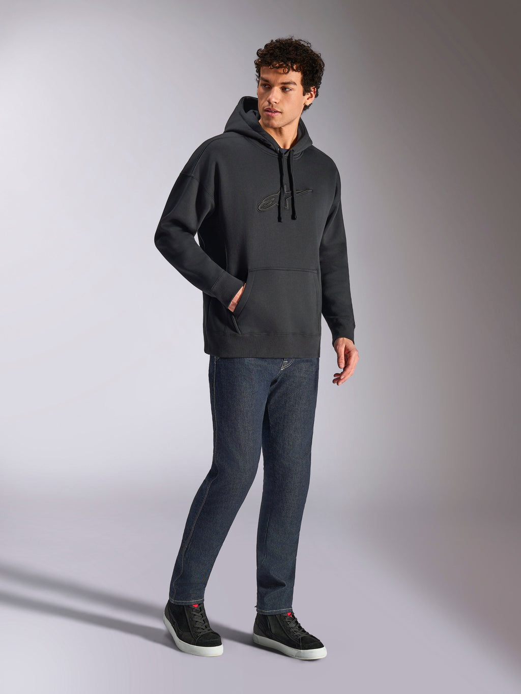 Alpinestars Emergent Oversized Hoodie, felpa casual streetwear nera, logo Astars ricamato in 3D tono su tono sul petto, tasca a marsupio, vestibilità oversize, indossata da un modello con jeans in denim blu scuro e sneakers nere
