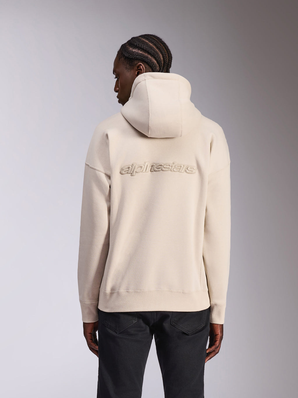 Alpinestars Emergent Oversized Hoodie, streetwear casual, colore bianco sporco Sand, logo Alpinestars ricamato in 3D sulla schiena, vestibilità oversized con orlo e polsini a costine, vista posteriore su modello con pantaloni neri