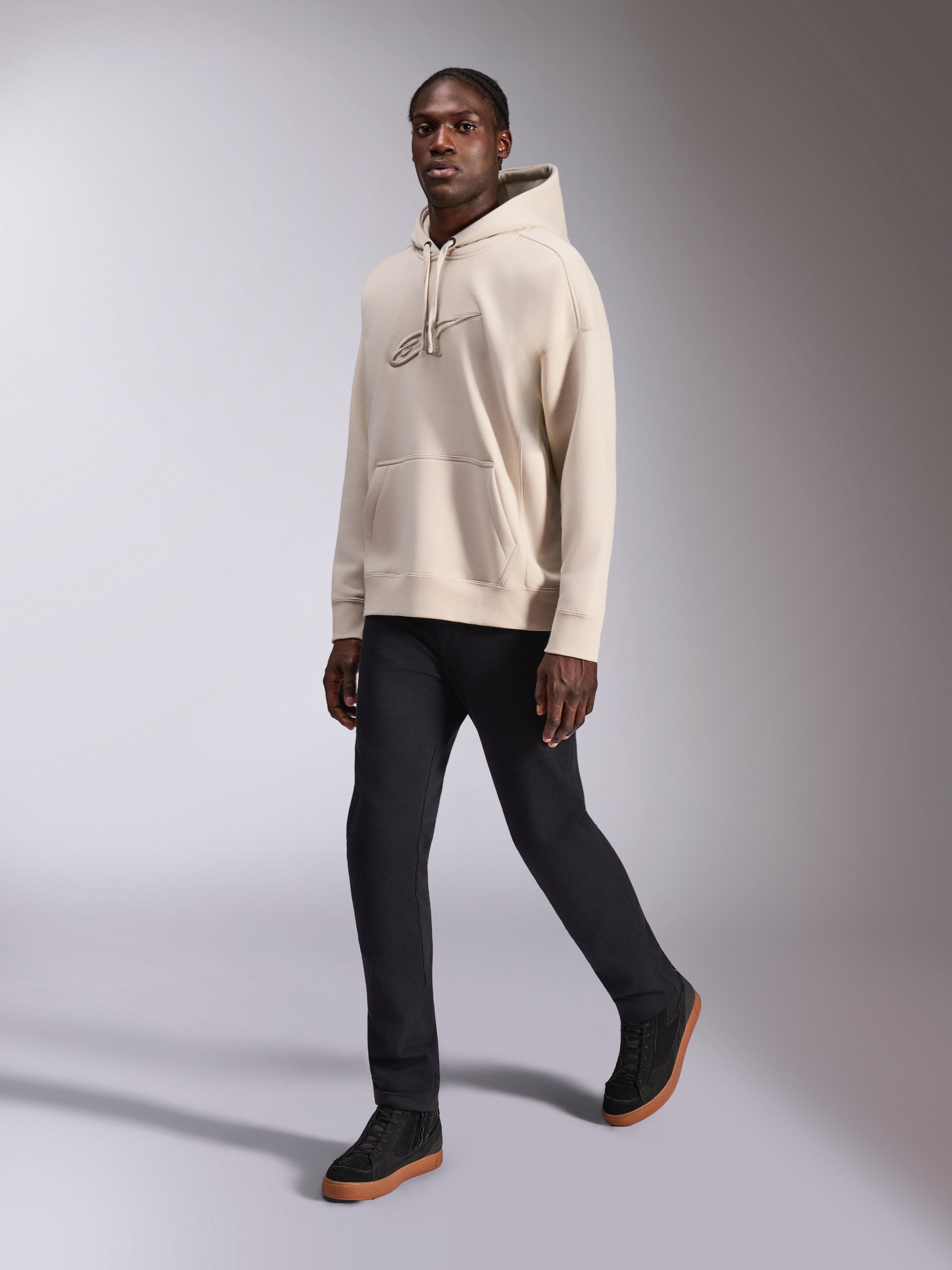 Alpinestars Emergent Oversized Hoodie, streetwear casual, bianco sporco sabbia, logo ricamato 3D in tinta, indossata da un modello con pantaloni neri e sneakers nere, vestibilità oversize rilassata con tasca a marsupio e cappuccio con coulisse
