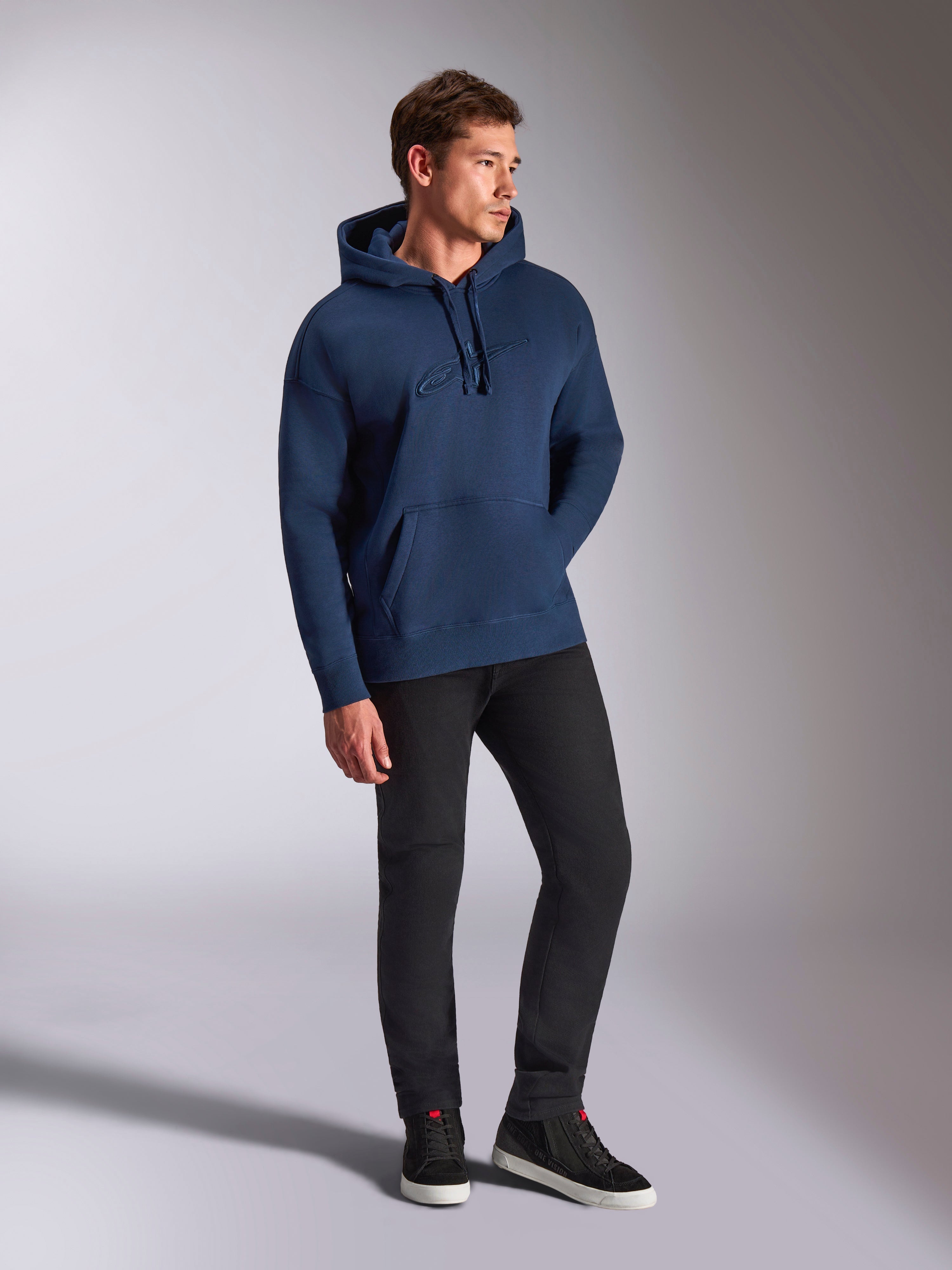 Felpa con cappuccio Alpinestars Emergent Oversized, blu navy, indossata da un modello con pantaloni neri e sneakers, con logo Astars ricamato in 3D, tasca a marsupio e cappuccio con cordino, streetwear casual ispirato al motorsport.