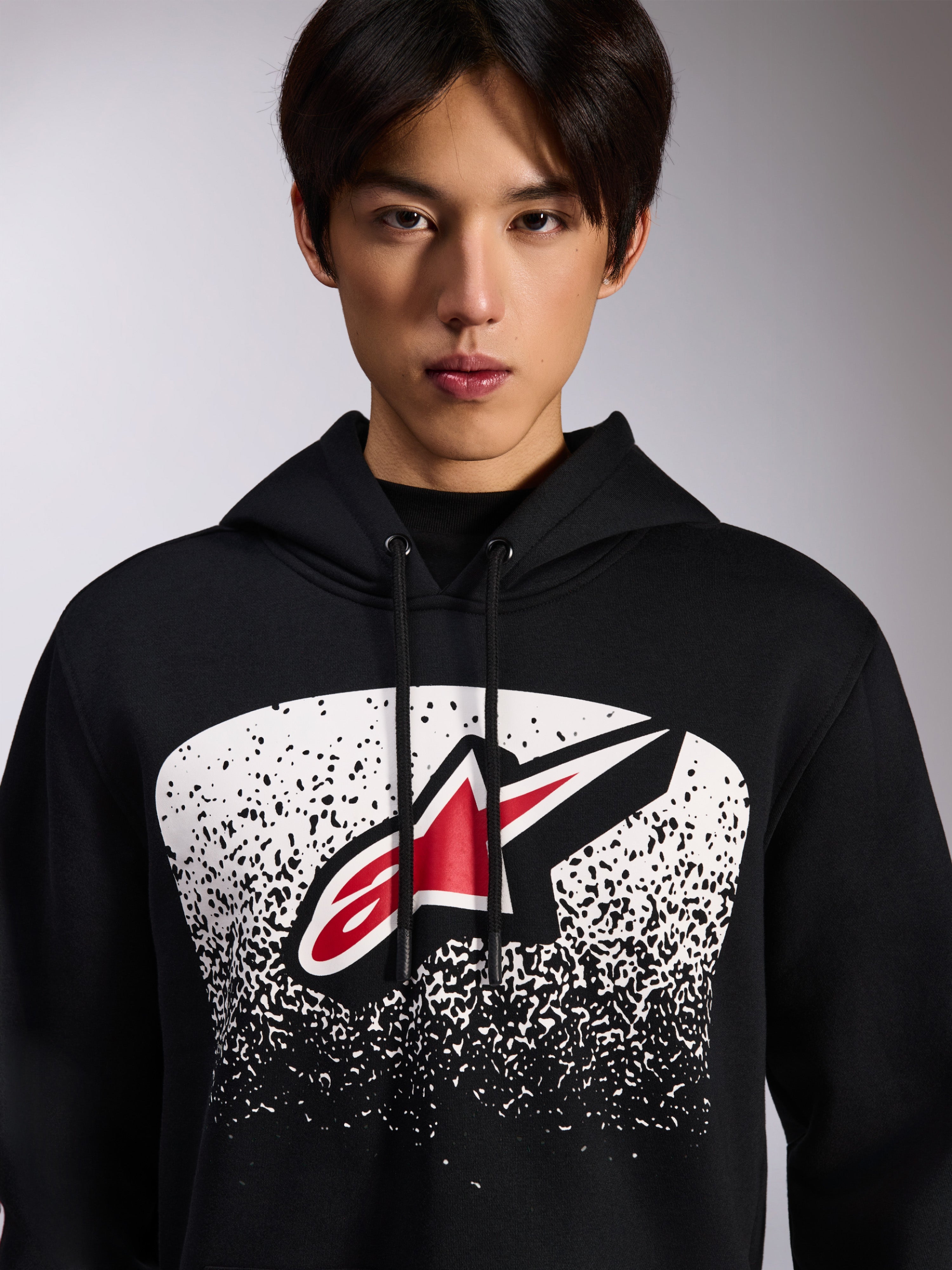 Alpinestars Obsessed Hoodie, felpa nera con cappuccio, con logo Astars rosso e bianco all'interno di una grafica bianca a gradazione puntinata MX sul petto, indossata da un modello, abbigliamento streetwear casual da motorsport con cappuccio regolabile con cordino