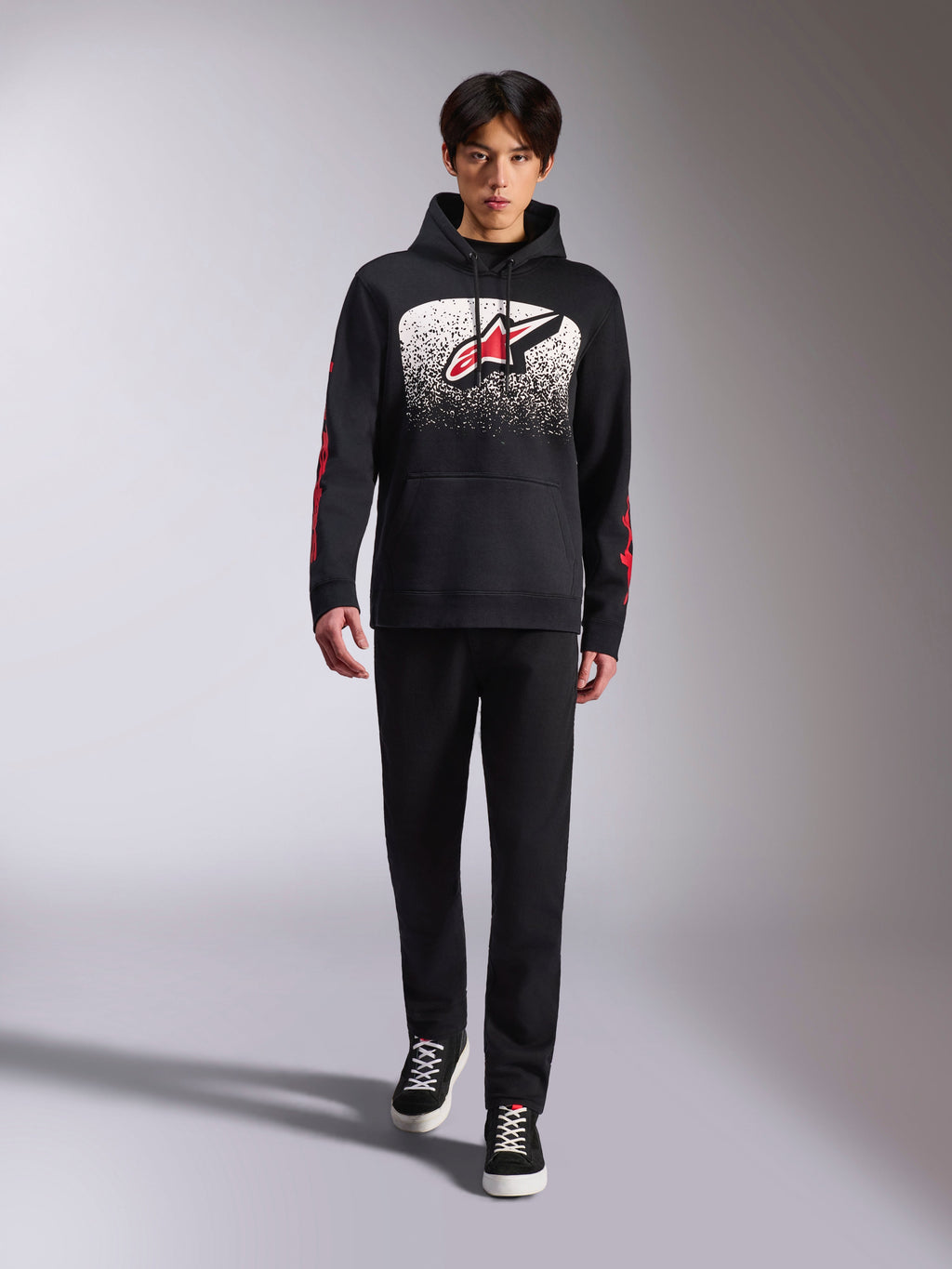 Alpinestars Obsessed Hoodie, felpa nera con cappuccio, grande logo Astars bianco e rosso con gradazione MX, scritte rosse sulle maniche, indossata da un modello con outfit casual total black, abbigliamento lifestyle ispirato al motorsport