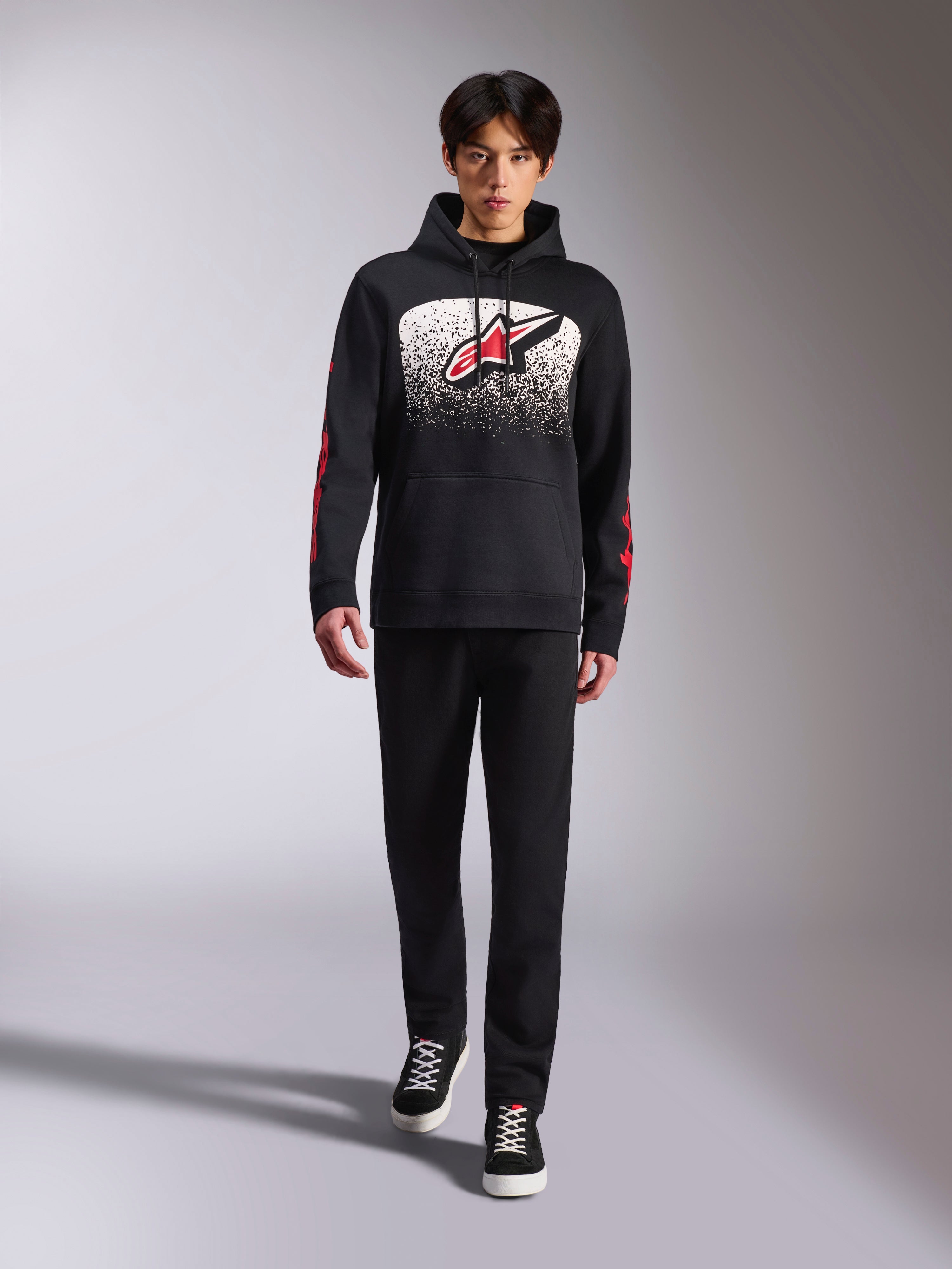 Alpinestars Obsessed Hoodie, felpa nera con cappuccio, grande logo Astars bianco e rosso con gradazione MX, scritte rosse sulle maniche, indossata da un modello con outfit casual total black, abbigliamento lifestyle ispirato al motorsport