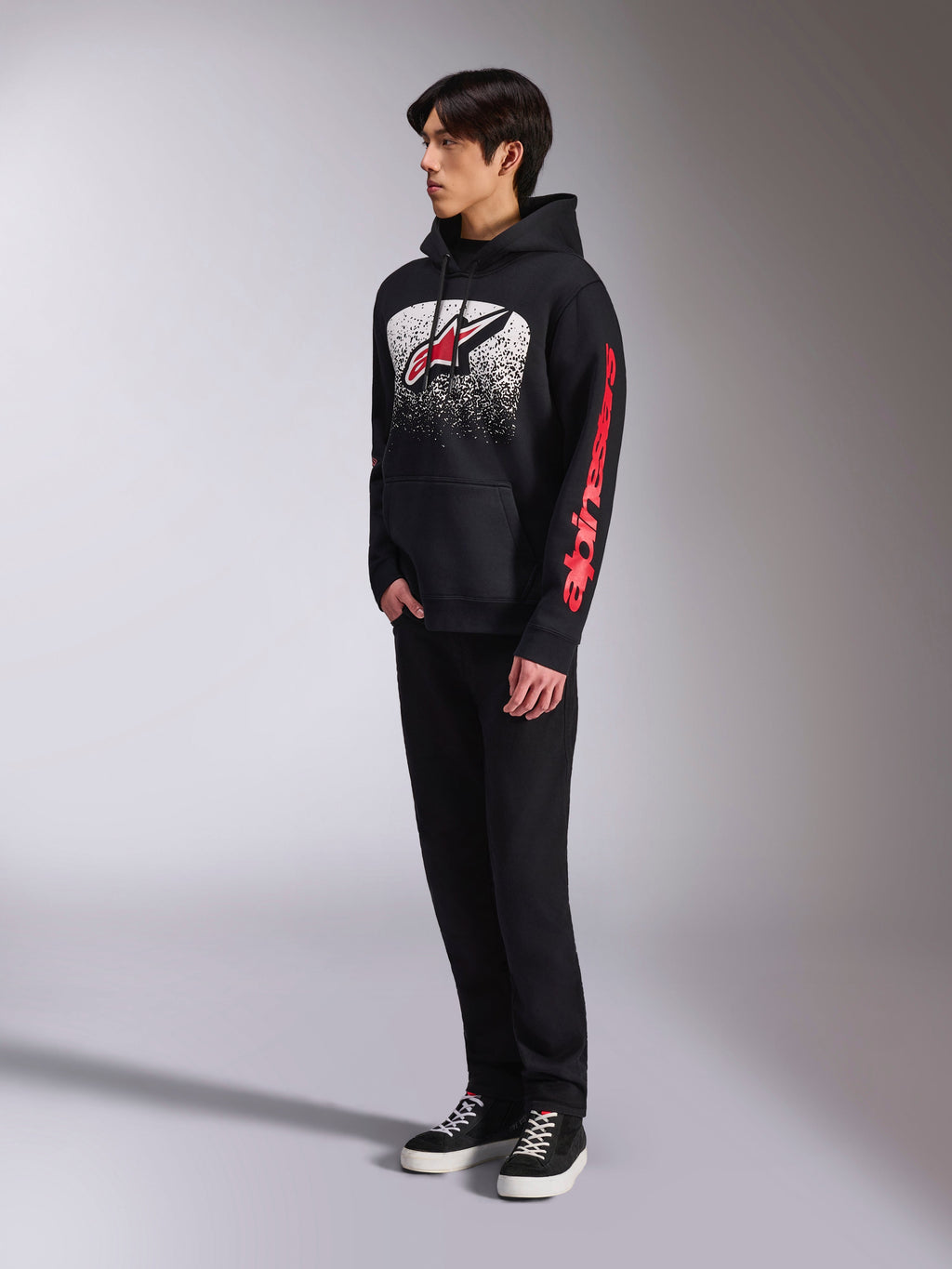 Alpinestars Obsessed Hoodie, felpa in pile nera, con una grande grafica bianca punteggiata sul petto, logo Astars rosso e branding rosso sulle maniche, indossata da un modello con pantaloni neri e sneaker, streetwear casual motorsport