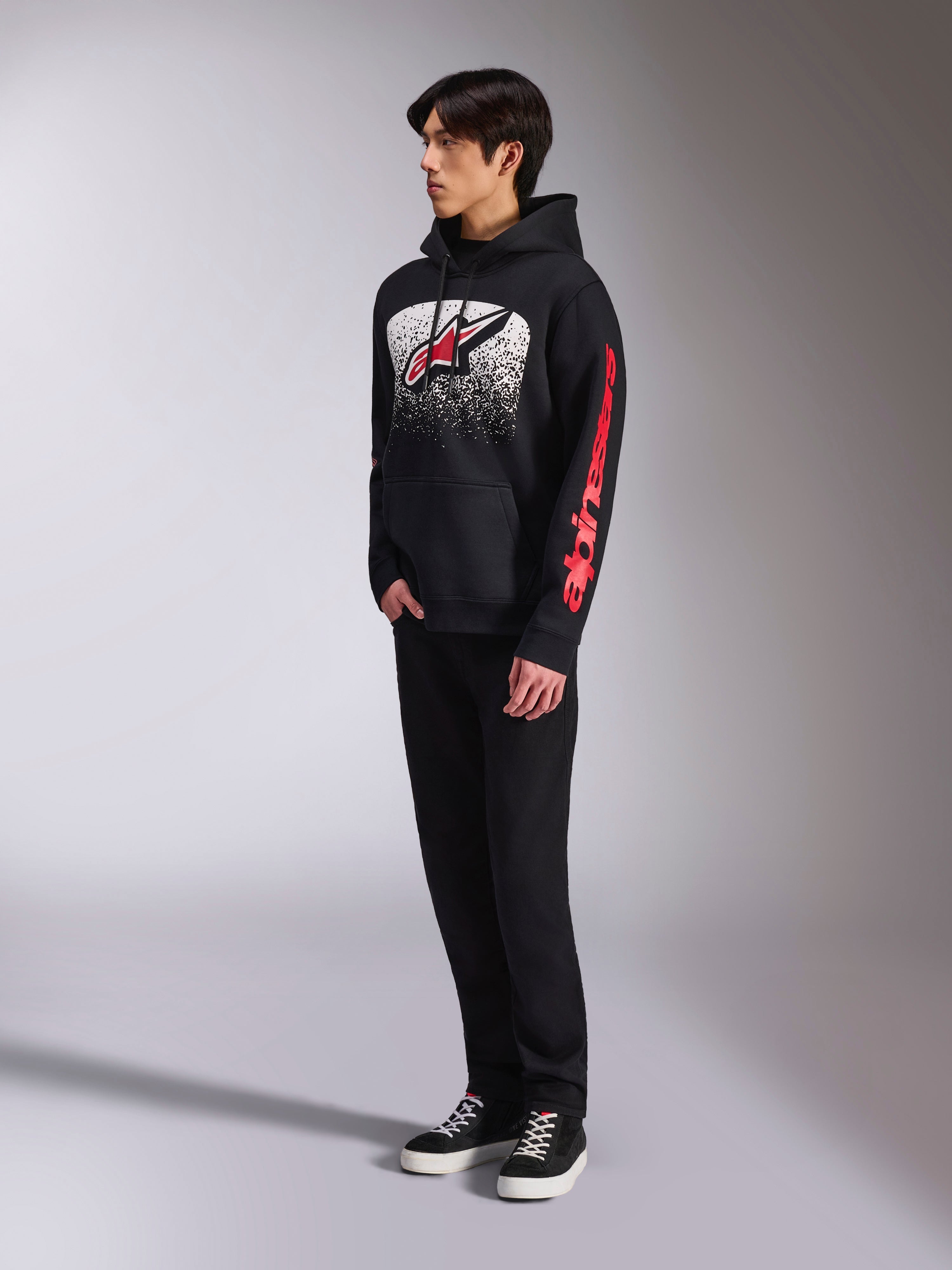 Alpinestars Obsessed Hoodie, felpa in pile nera, con una grande grafica bianca punteggiata sul petto, logo Astars rosso e branding rosso sulle maniche, indossata da un modello con pantaloni neri e sneaker, streetwear casual motorsport