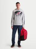 Alpinestars Felpa con cappuccio Obsessed, felpa sportswear da uomo in pile, grigio cemento, grafiche MX sfumate rosse e nere sul petto, branding sulle maniche, indossata da un modello con uno zaino rosso Alpinestars.