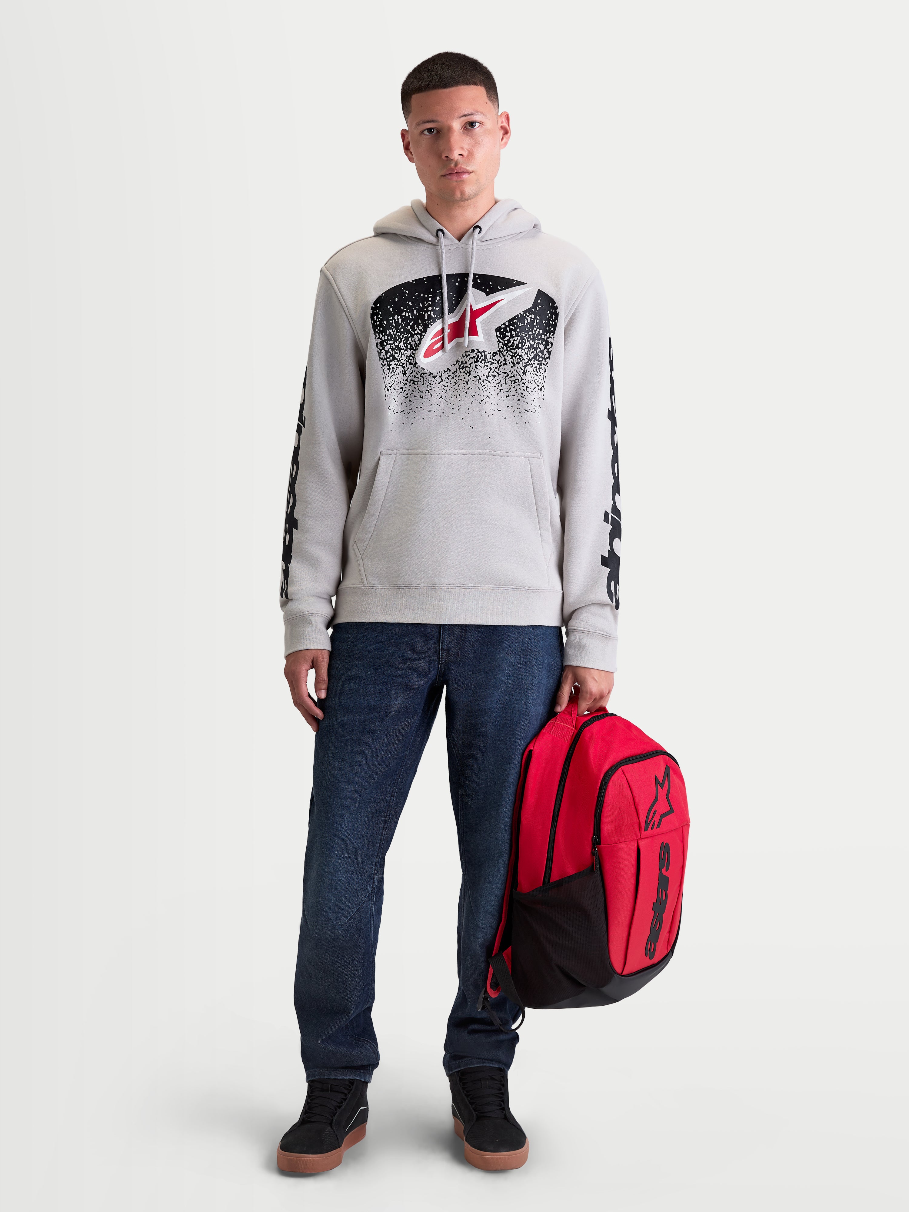 Alpinestars Felpa con cappuccio Obsessed, felpa sportswear da uomo in pile, grigio cemento, grafiche MX sfumate rosse e nere sul petto, branding sulle maniche, indossata da un modello con uno zaino rosso Alpinestars.
