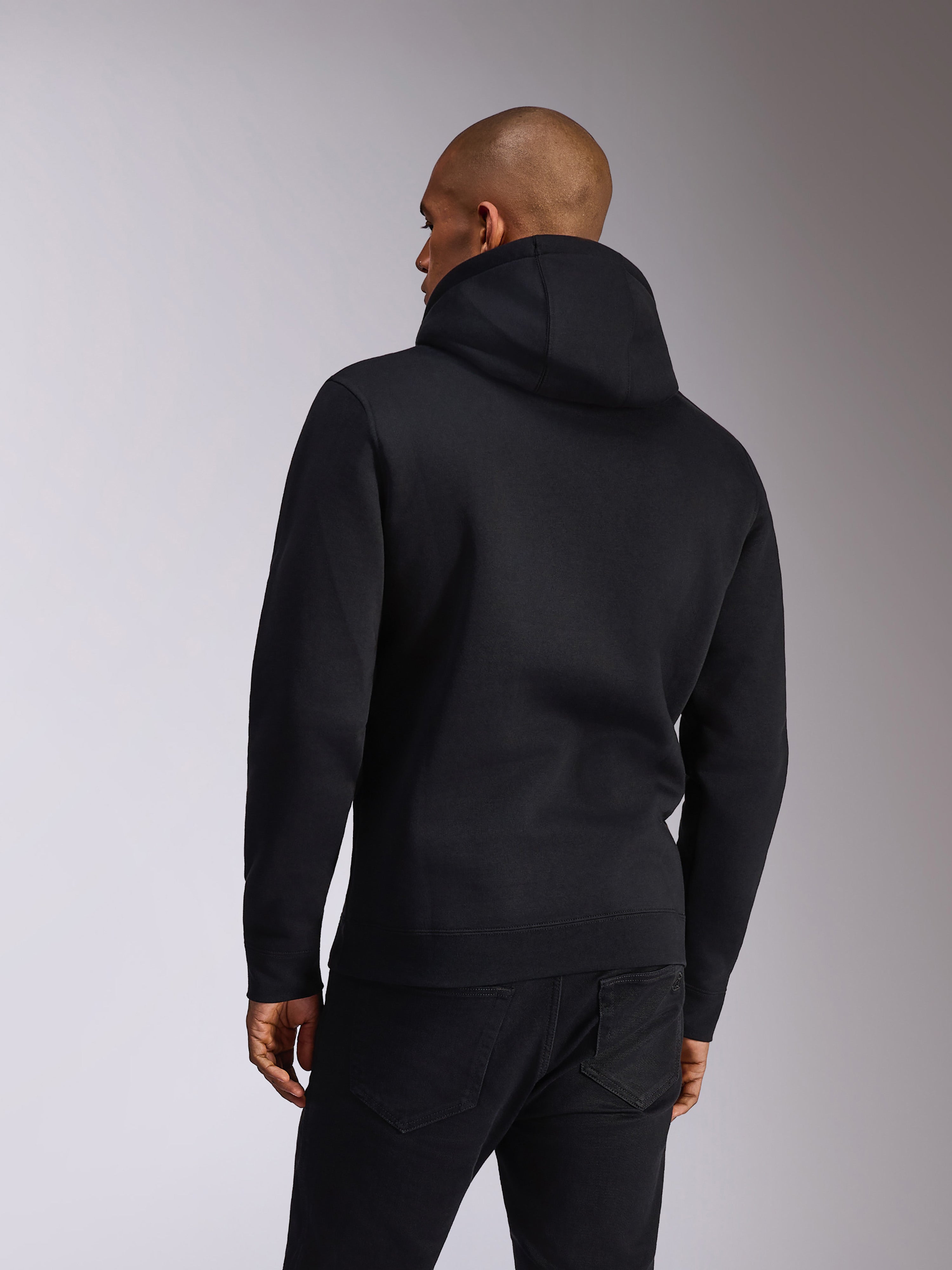 Alpinestars Ellipsoid Hoodie, nero, vista posteriore di una persona che indossa una felpa pullover con cappuccio, maniche lunghe con polsini e orlo a coste, abbinata a jeans neri, abbigliamento sportivo casual ispirato al motorsport